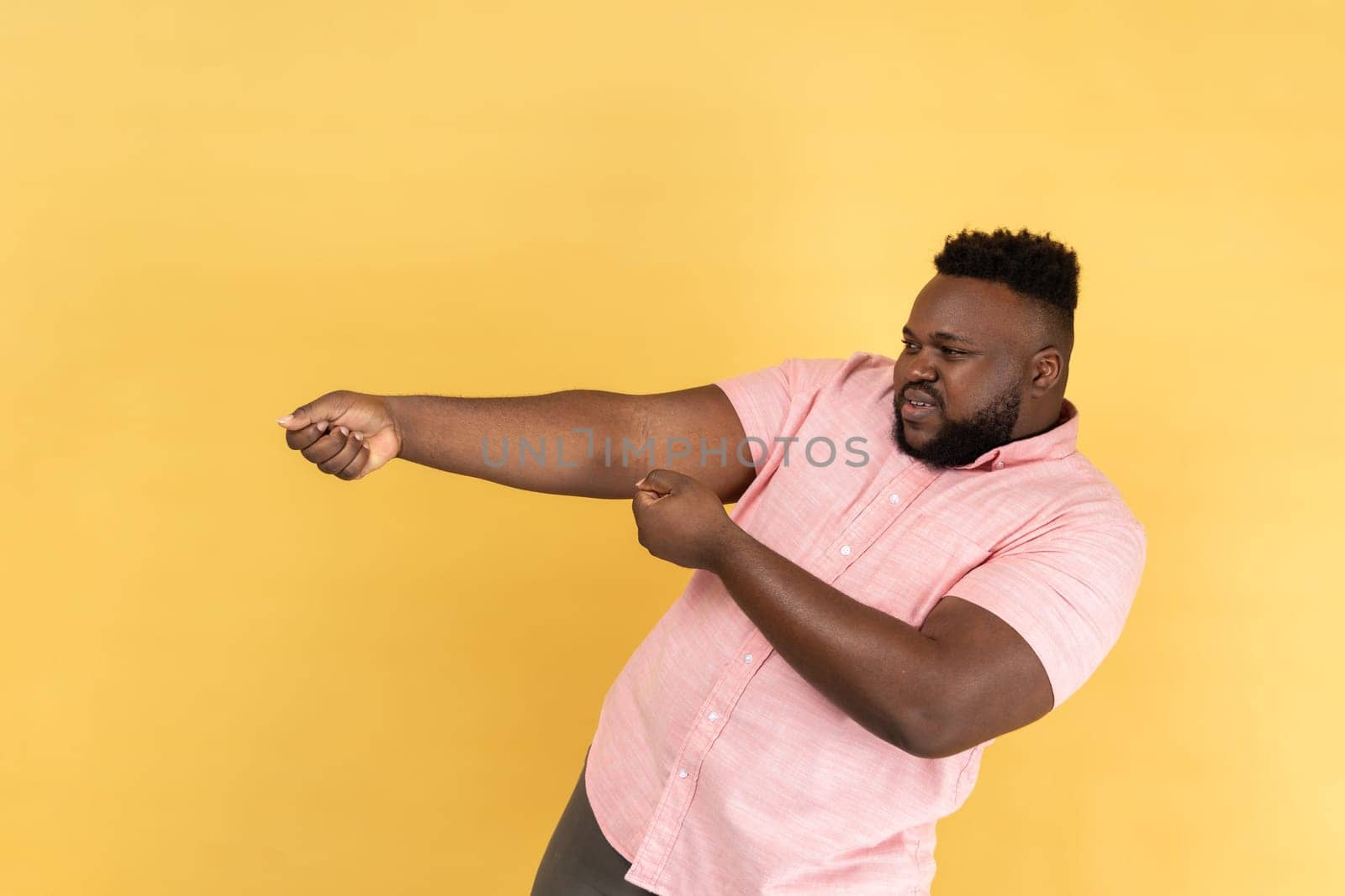 Portrait of persistent man with beard wearing pink shirt pretending to pull, , holding heavy virtual burden with expression of great effort. Indoor studio shot isolated on yellow background.