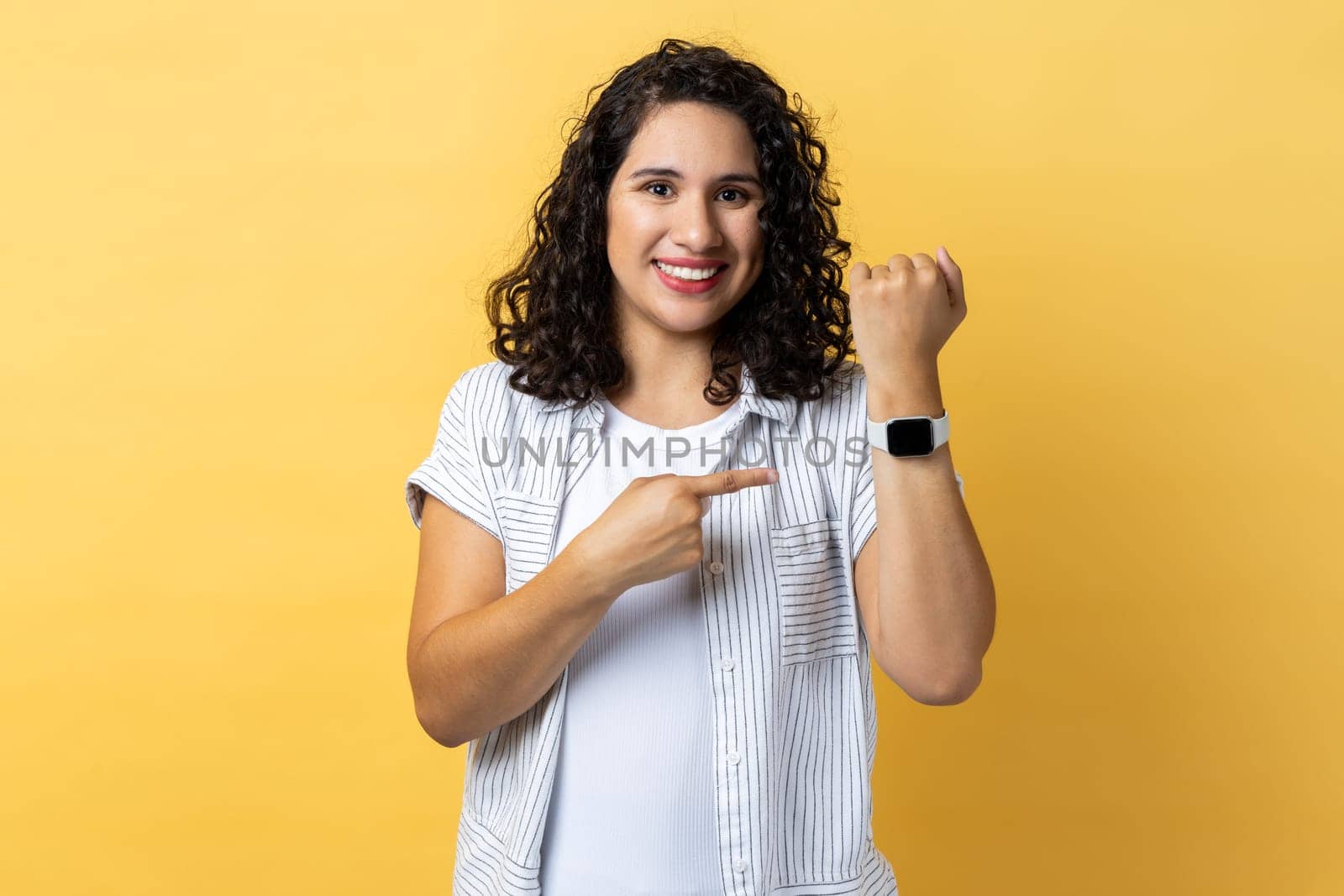 Portrait of positive woman with dark wavy hair standing, looking at camera, pointing at her smart watch, expressing happiness. Indoor studio shot isolated on yellow background.