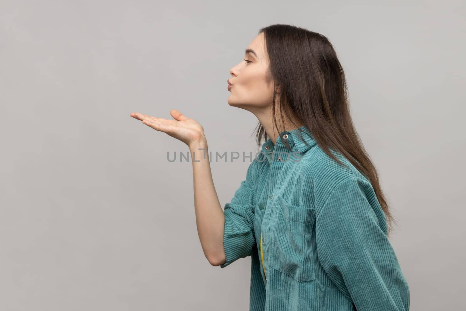 Catch my kiss. Side view of lovely romantic young woman looking ahead air kissing over palms hands, sending love, wearing casual style jacket. Indoor studio shot isolated on gray background.