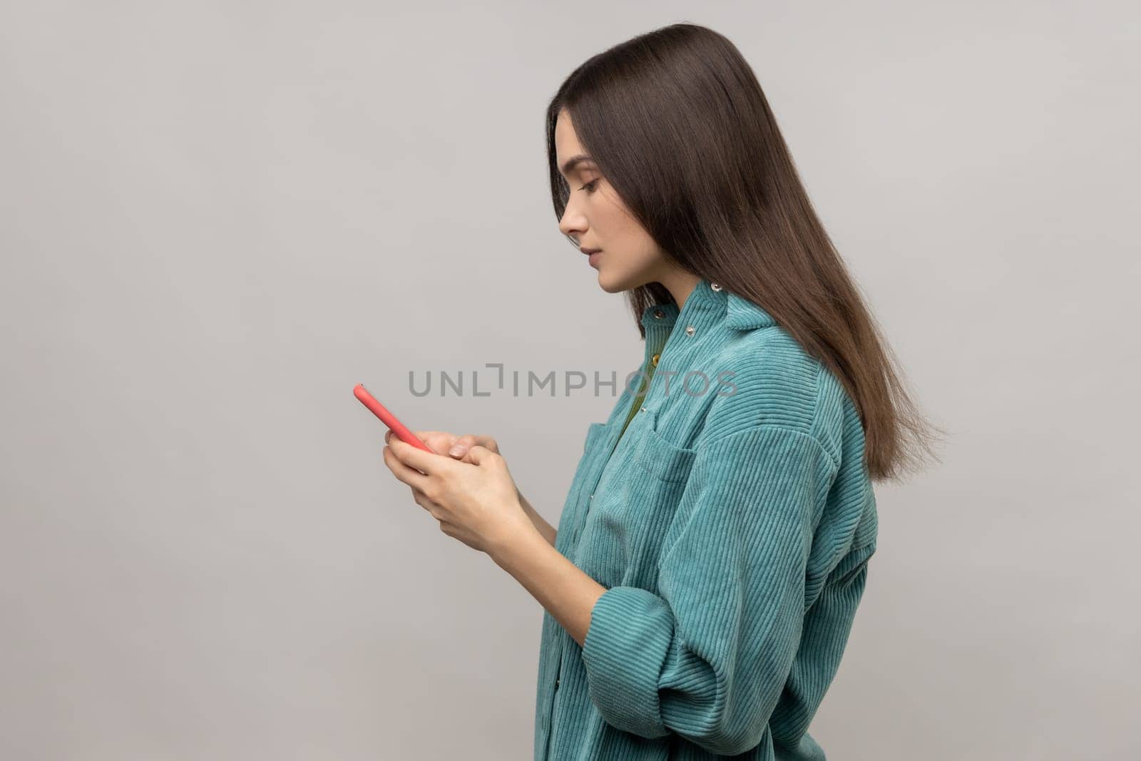 Side view of serious pretty woman reading message on smartphone, using mobile device for communication, browsing web, wearing casual style jacket. Indoor studio shot isolated on gray background.