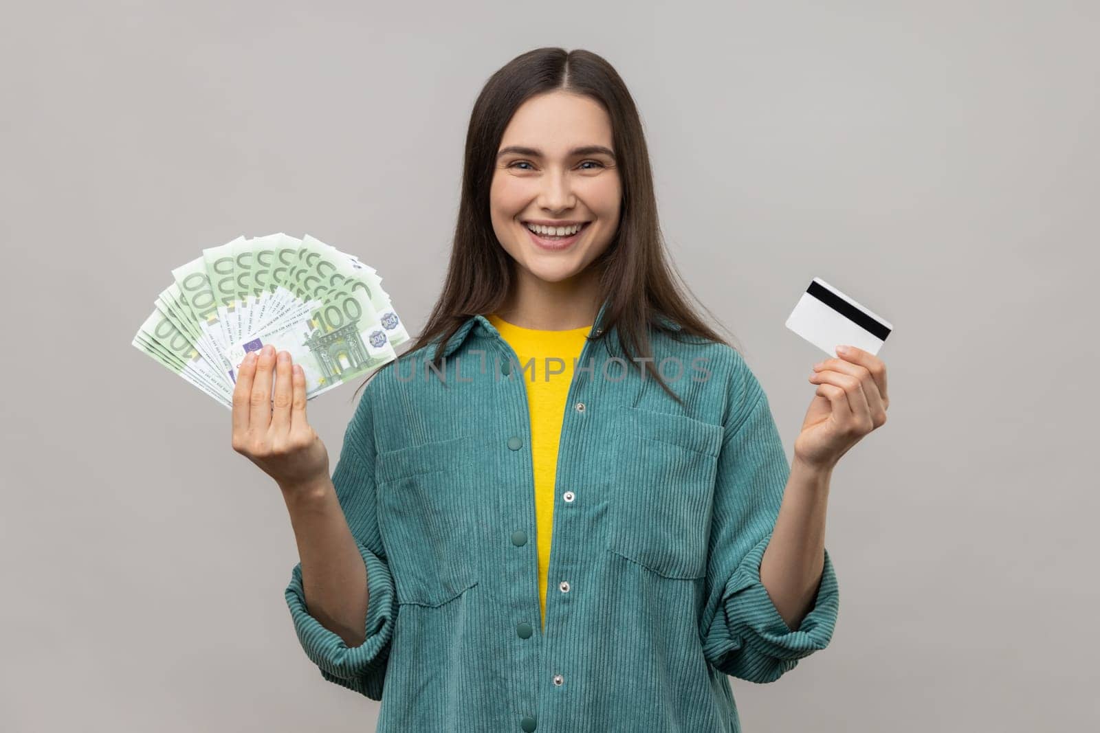 Happy satisfied young woman holding credit card and fan of euro cash looking at camera with toothy smile, wearing casual style jacket. Indoor studio shot isolated on gray background.