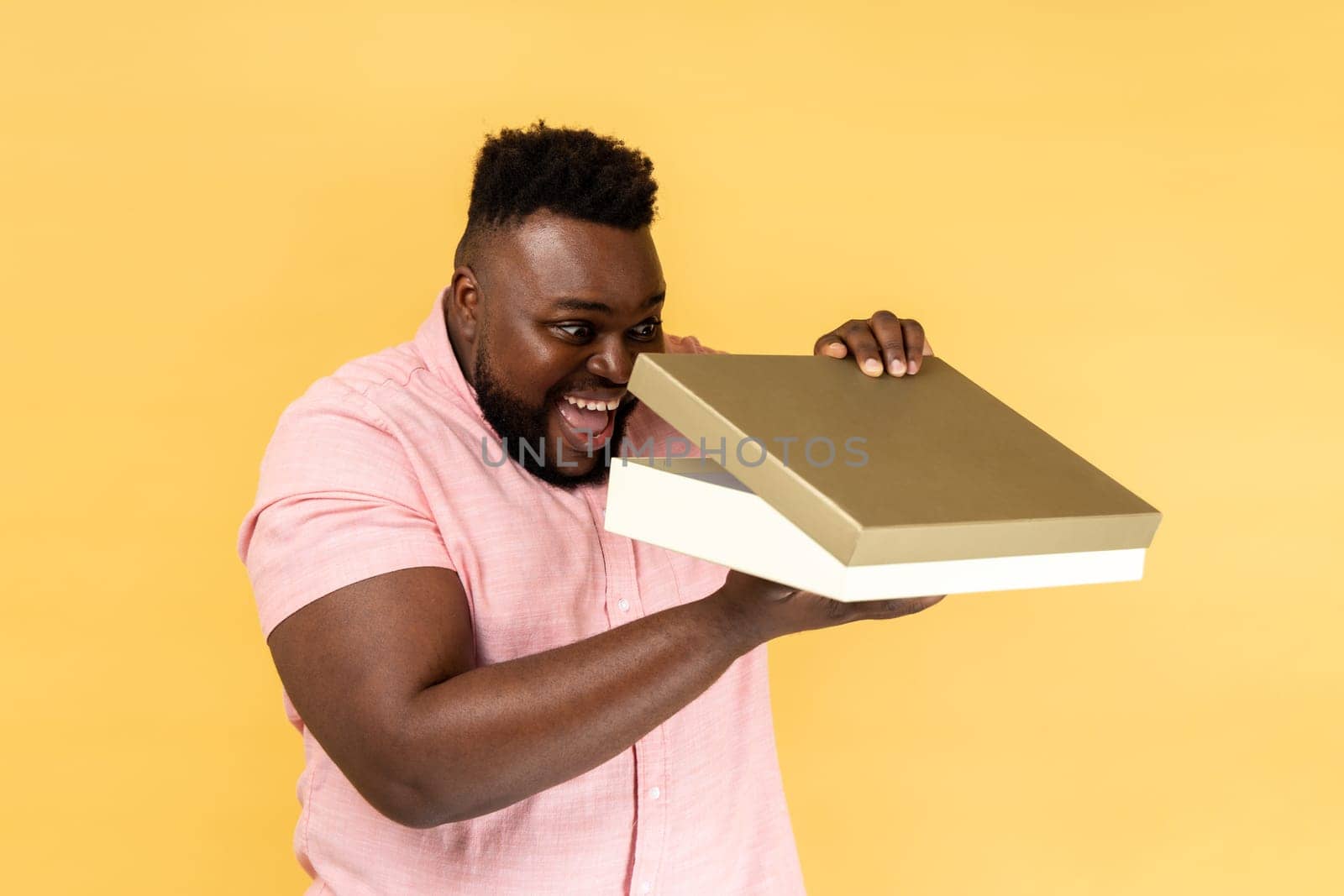 Portrait of optimistic man wearing pink shirt opening gift box and peeping inside with toothy smile, satisfied with present. Indoor studio shot isolated on yellow background.