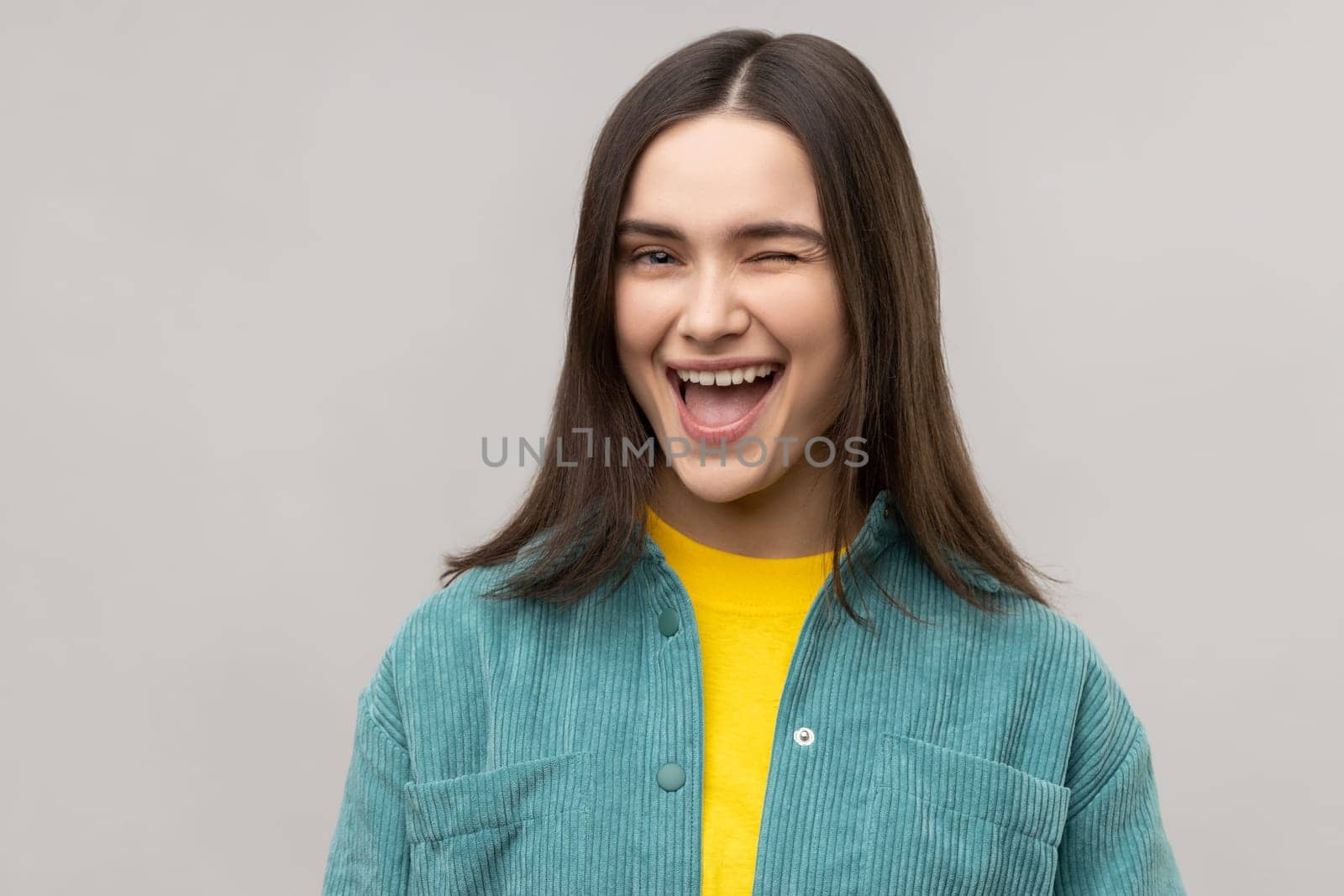 Portrait of good looking cheerful woman winking eye and flirting with someone, looks at camera, expressing happiness, wearing casual style jacket. Indoor studio shot isolated on gray background.