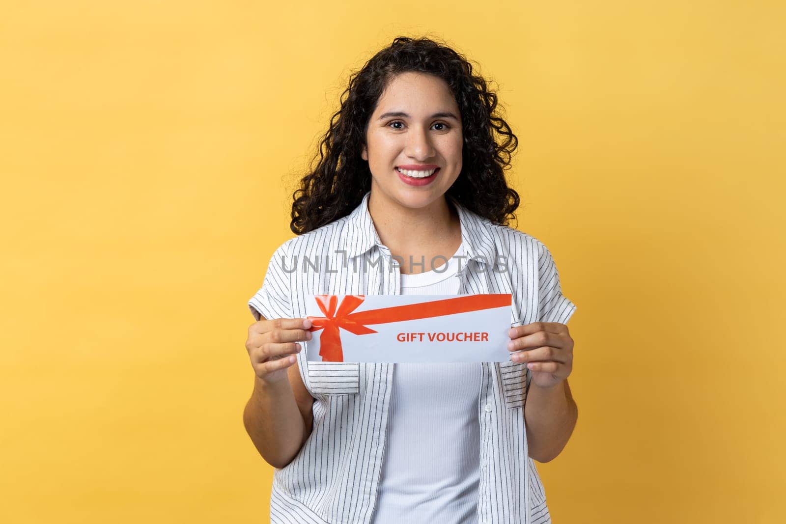 Portrait of delighted happy satisfied woman with dark wavy hair holding gift voucher, holiday gift card, coupon and certificate. Indoor studio shot isolated on yellow background.