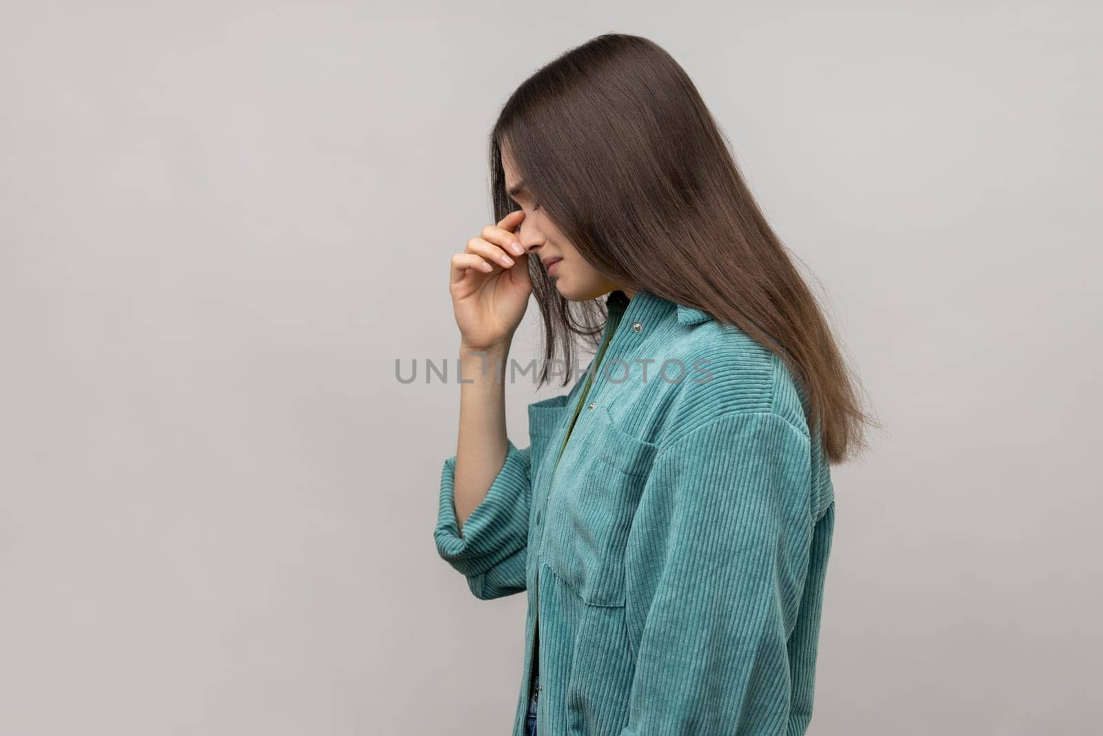 Side view of depressed woman holding her head down, touching face and crying with dramatic grimace, feeling sorrow regret, wearing casual style jacket. Indoor studio shot isolated on gray background.