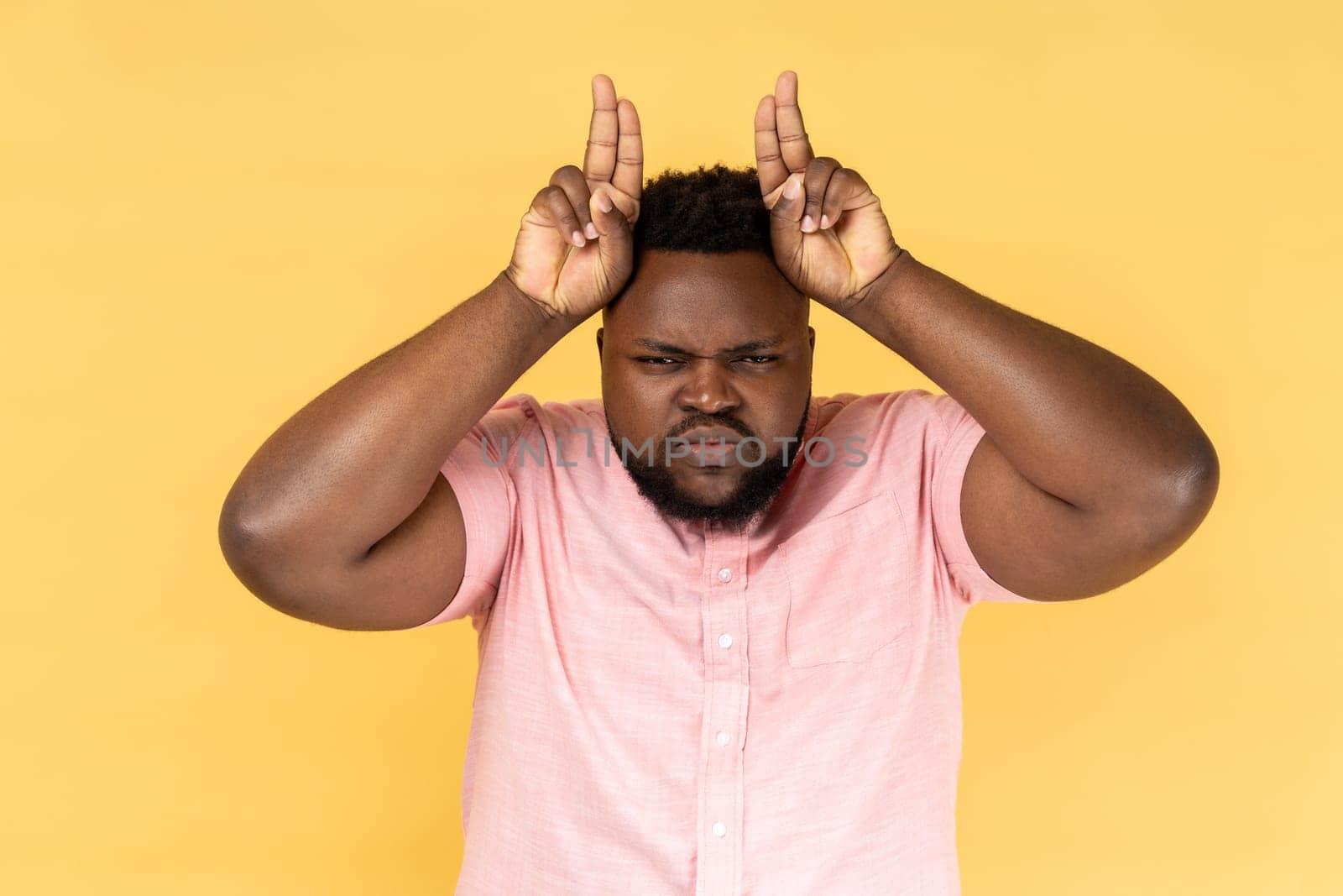 Portrait of man wearing pink shirt showing bull horn gesture, looking at camera with hostile angry expression, bully male threatening to attack. Indoor studio shot isolated on yellow background.