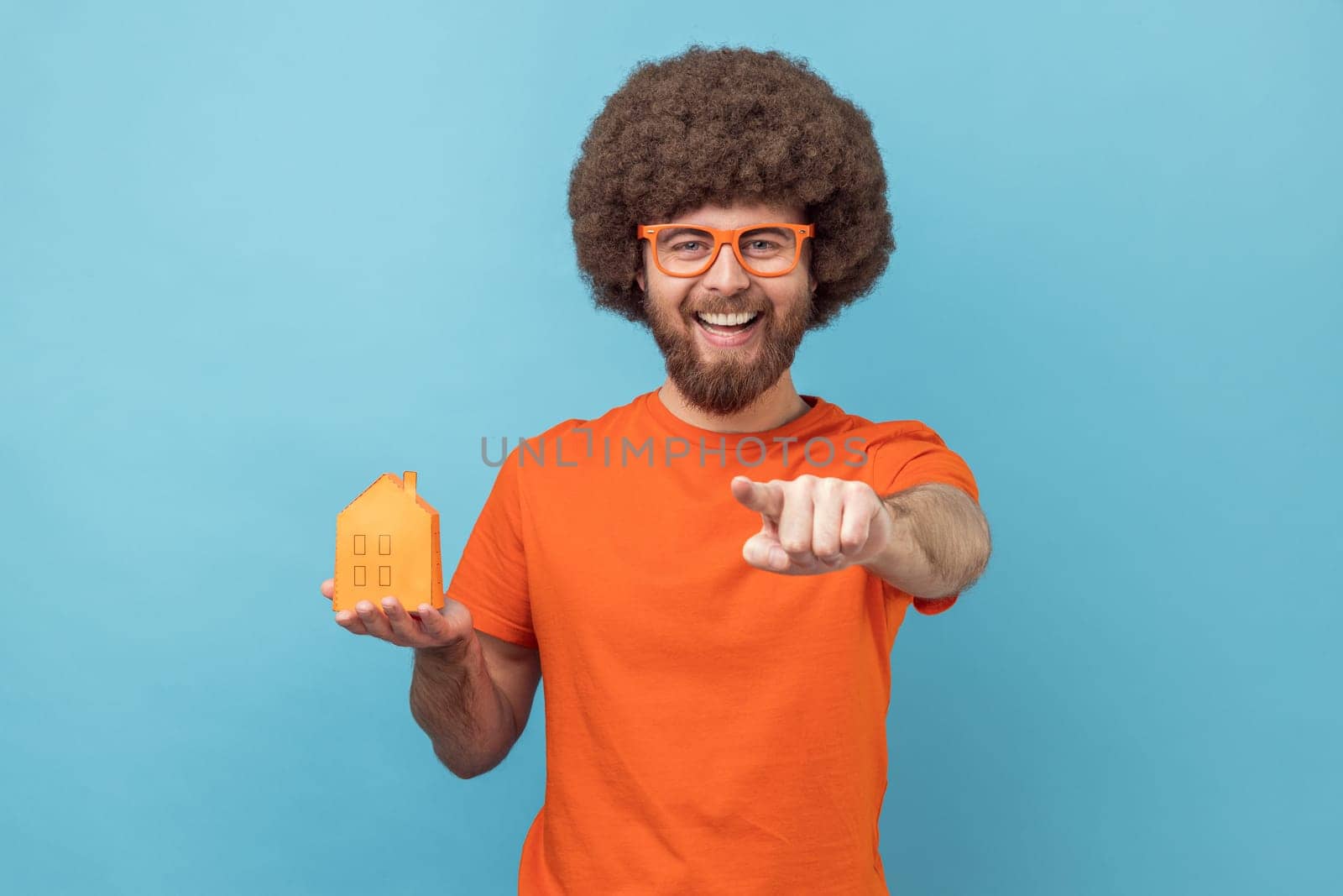 Satisfied man with Afro hairstyle wearing orange T-shirt holding paper house in hands, pointing at camera, can help you with buying real estate. Indoor studio shot isolated on blue background.