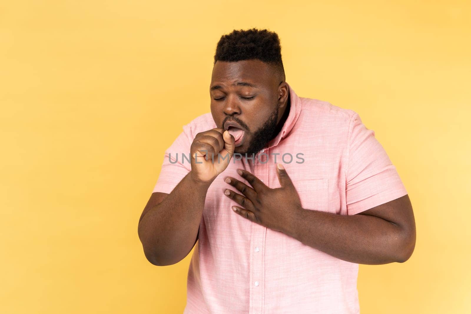 Portrait of sick unhealthy bearded man wearing pink shirt having cough and feeling pain in her lungs, catching cold, flu symptoms. Indoor studio shot isolated on yellow background.
