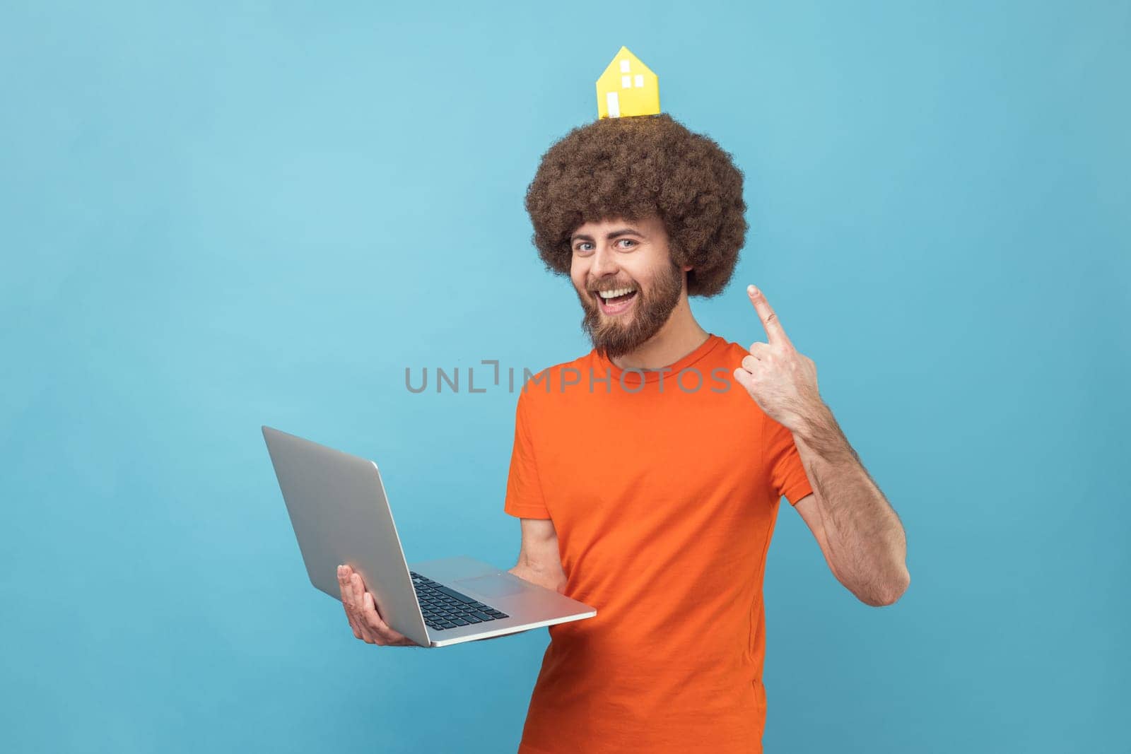 Portrait of funny man with Afro hairstyle wearing orange T-shirt holding notebook and pointing at paper house on his head, real estate. Indoor studio shot isolated on blue background.