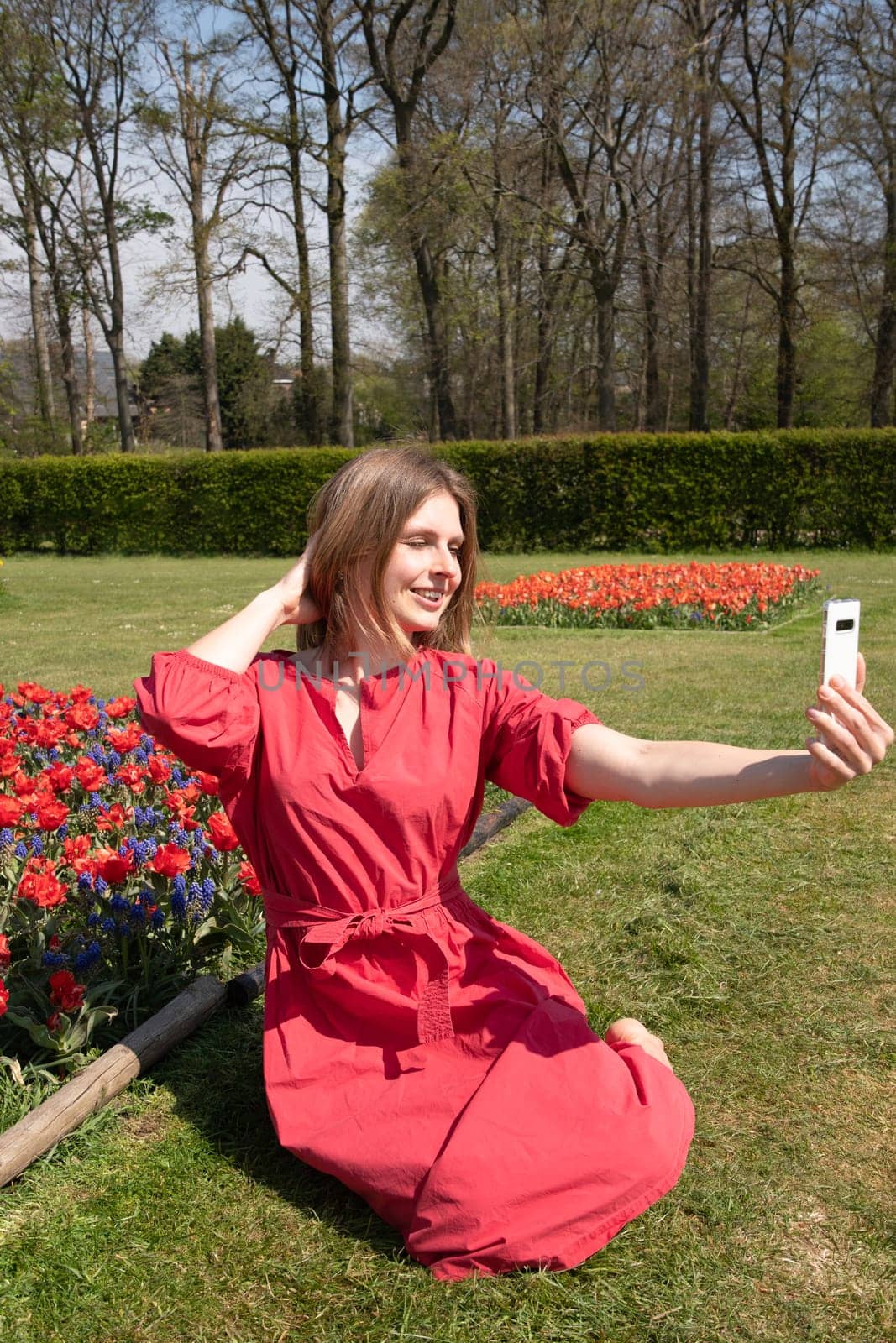 A young beautiful girl in a red dress in a spring park plays and takes a selfie by KaterinaDalemans
