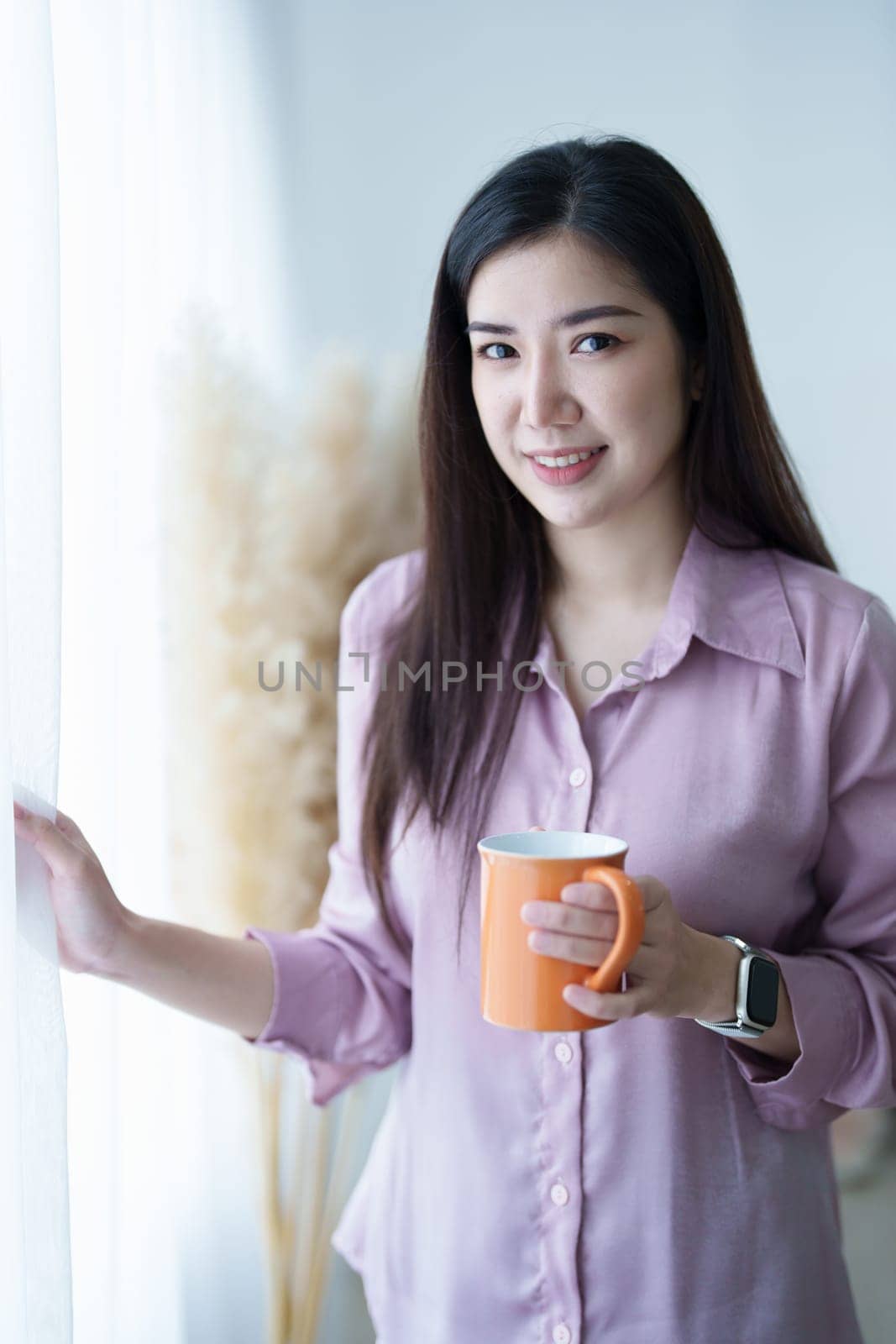 A portrait of a young Asian woman in pajamas smiling happily by a window with curtains in the morning sun while drinking coffee.