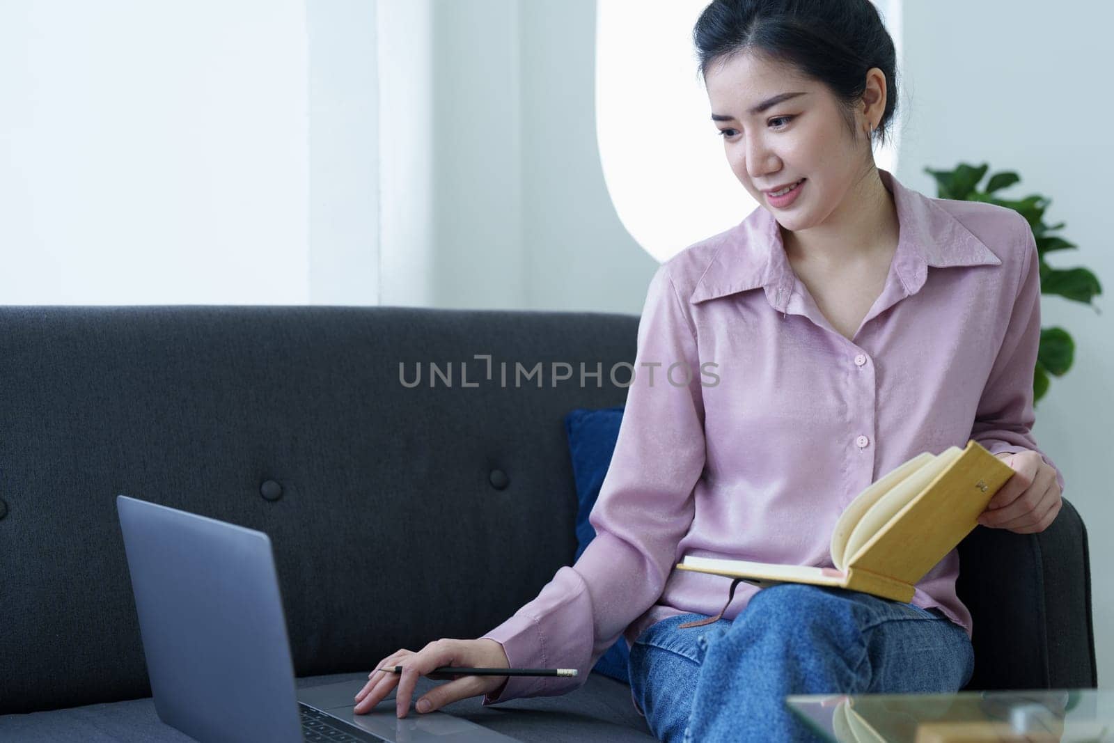 Portrait of a self-employed Asian woman holding a notebook and using a computer doing video conference at home by Manastrong