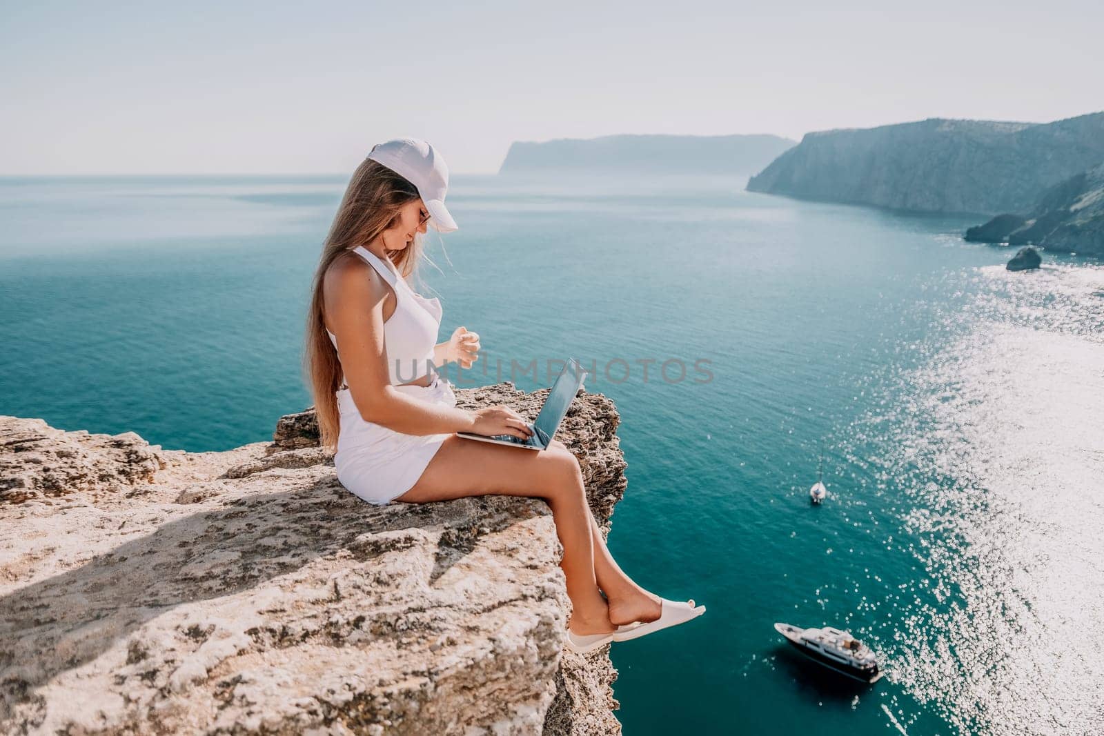 Successful business woman in yellow hat working on laptop by the sea. Pretty lady typing on computer at summer day outdoors. Freelance, travel and holidays concept.