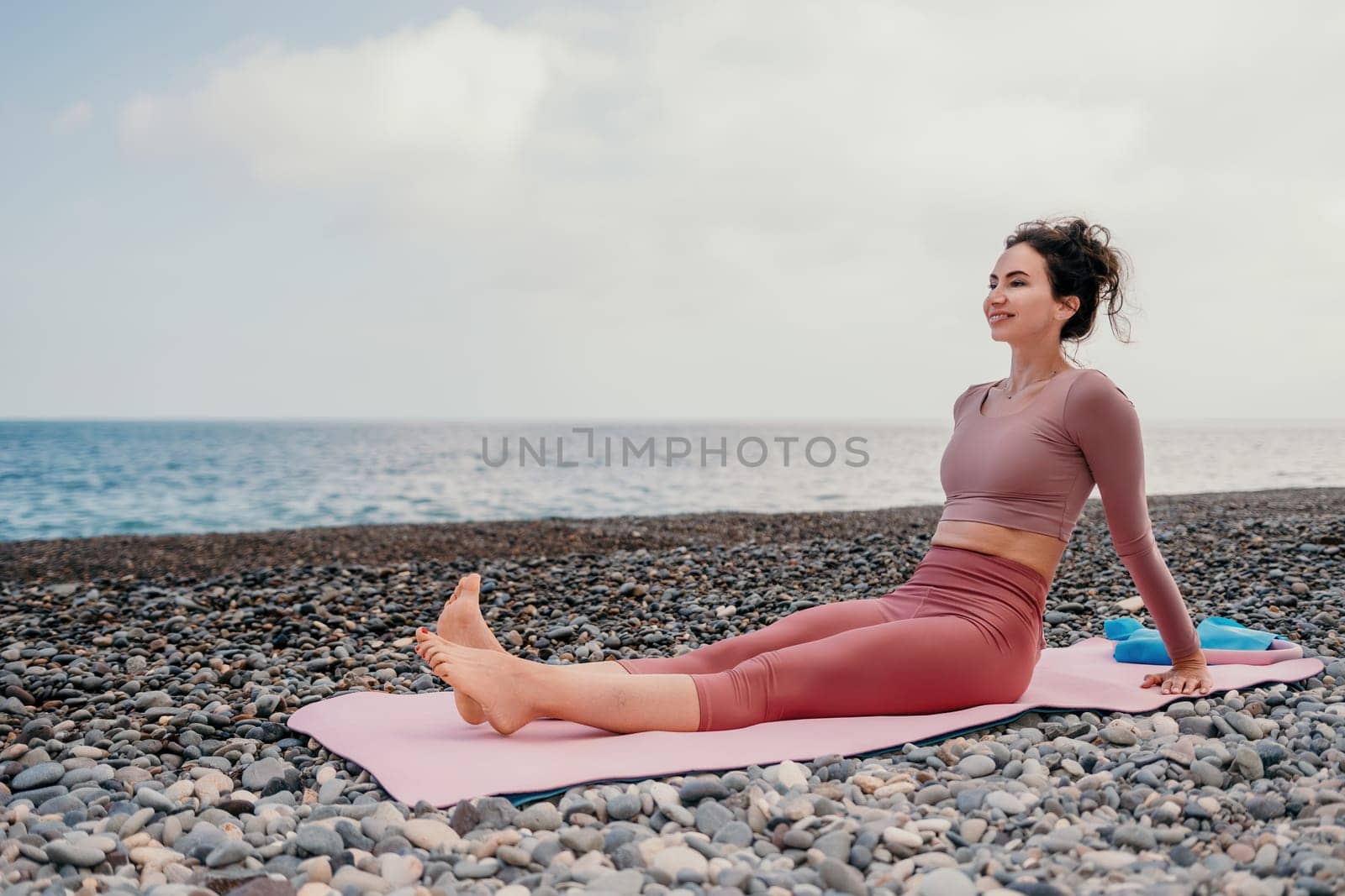 Middle aged well looking woman with black hair doing Pilates with the ring on the yoga mat near the sea on the pebble beach. Female fitness yoga concept. Healthy lifestyle, harmony and meditation.