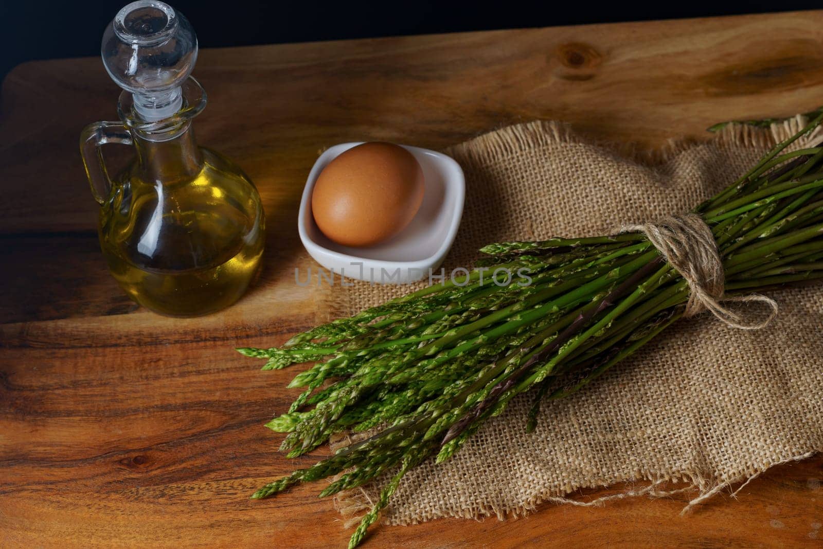 bunch of wild asparagus on a raffia cloth with a jar of olive oil and eggs black background
