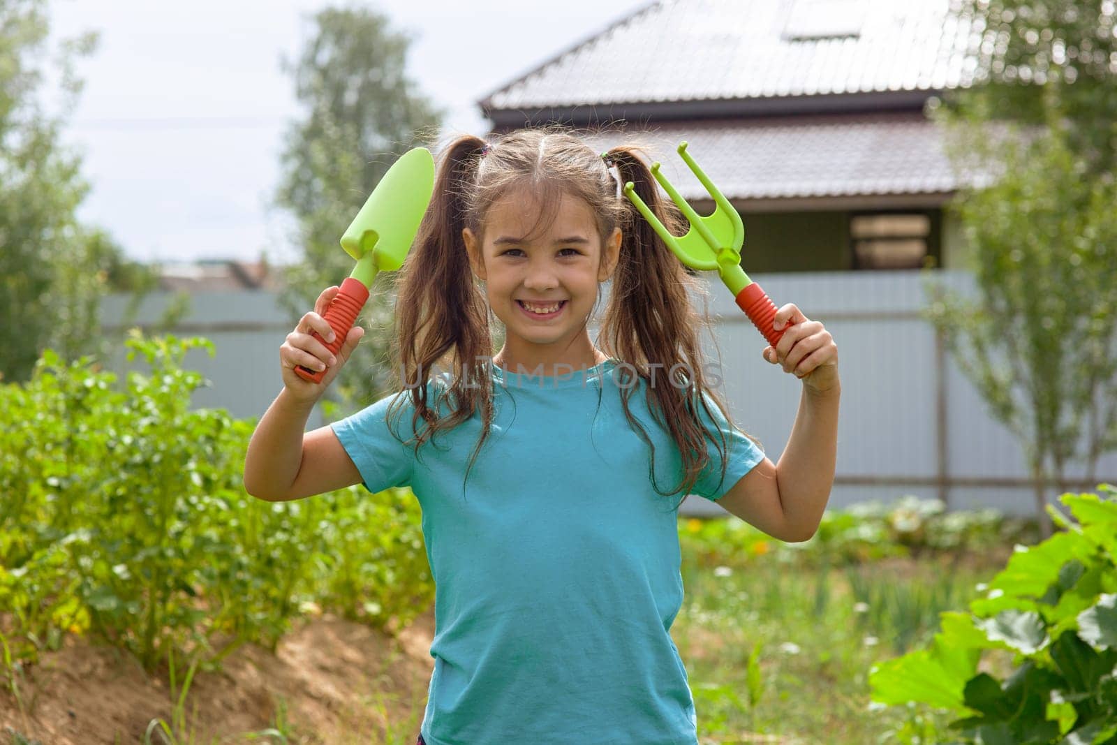 Cute little girl in green t-shirt holding small gardening tools , standing in the garden on a sunny day. Copy space