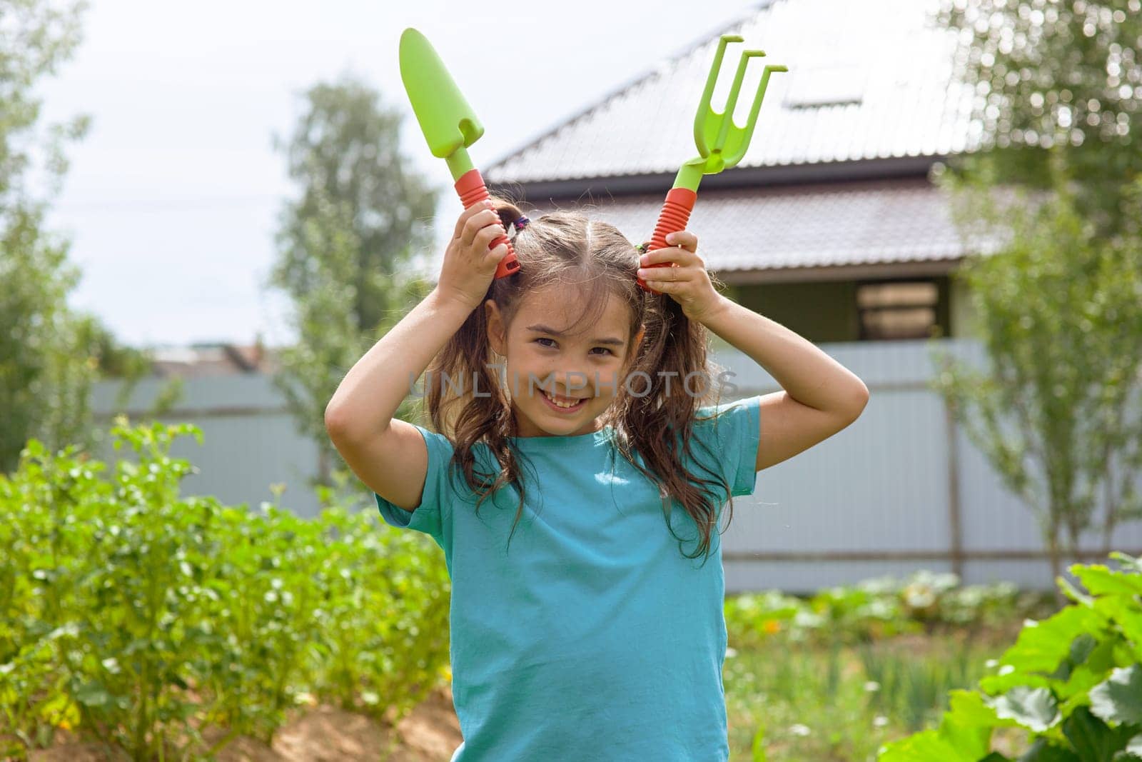 Funny little girl holding small gardening tools , standing in the garden on a sunny day. Copy space