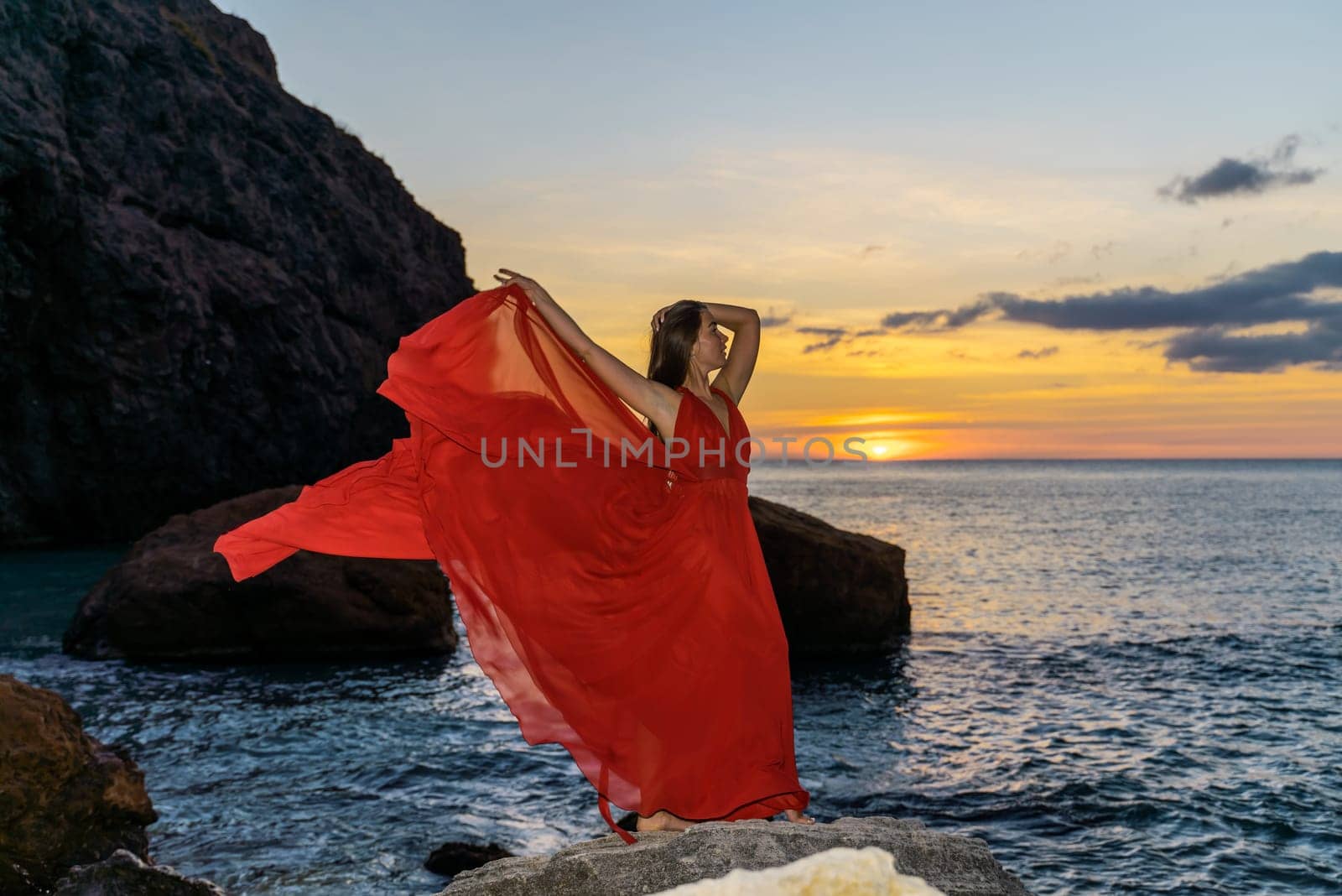 Woman in a red flying dress on the ocean or on the sea beach against the backdrop of the sunset sky. by Matiunina