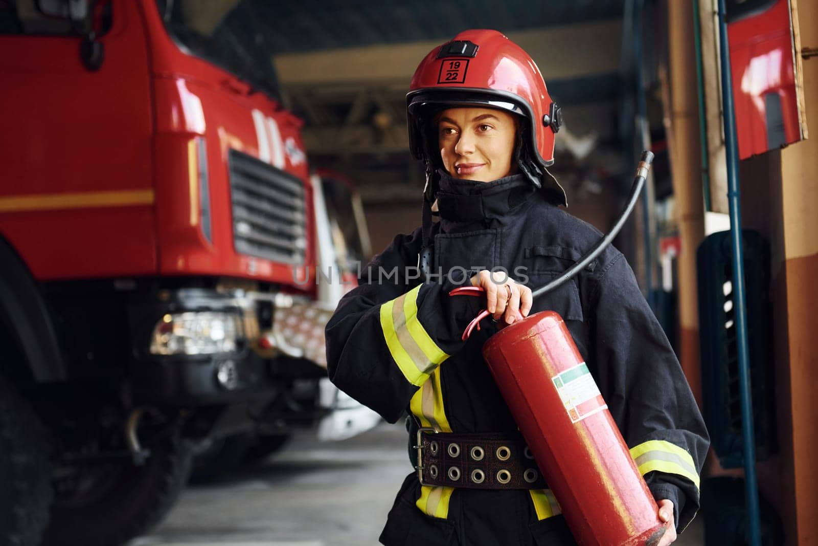 Holds extinguisher in hands. Female firefighter in protective uniform standing near truck.