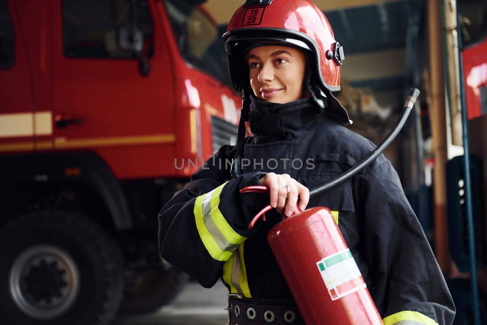 Holds extinguisher in hands. Female firefighter in protective uniform standing near truck.