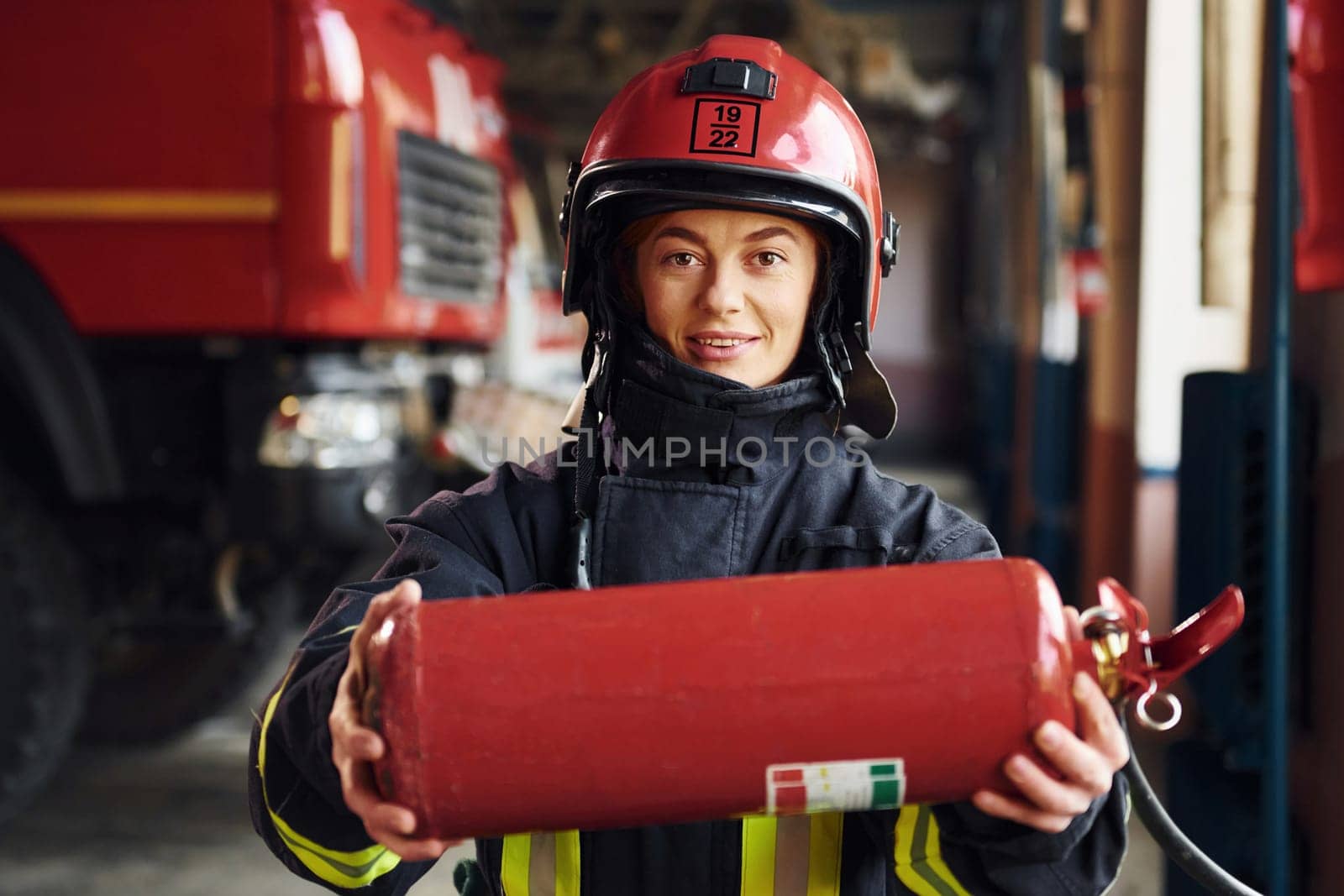 Holds extinguisher in hands. Female firefighter in protective uniform standing near truck.