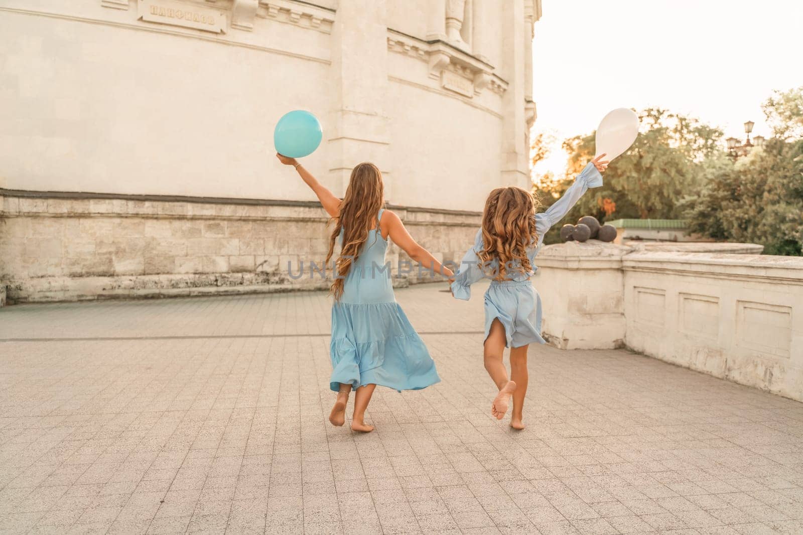 Daughter mother run holding hands. In blue dresses with flowing long hair, they hold balloons in their hands against the backdrop of a sunset and a white building. by Matiunina