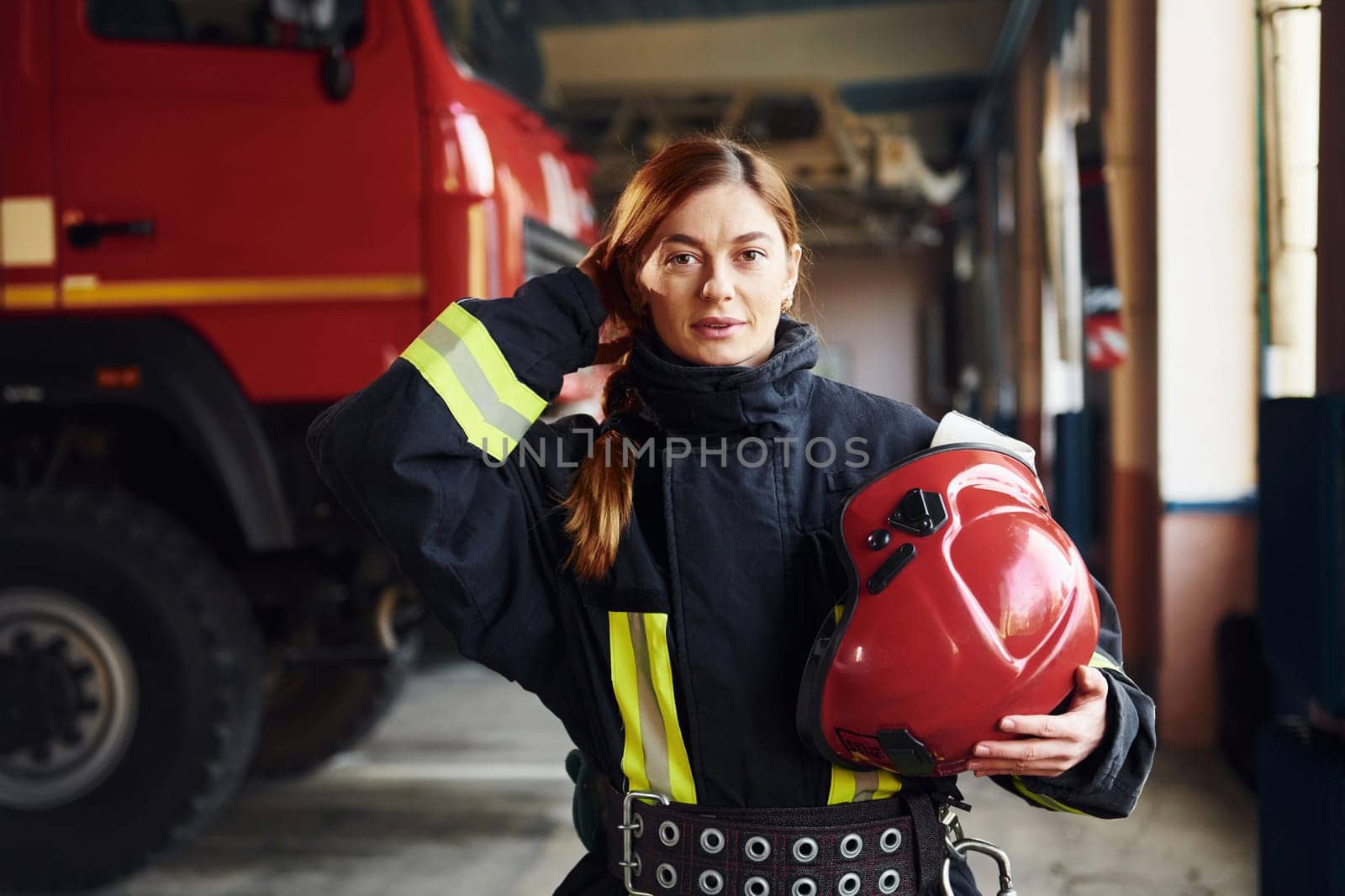 Holds red hat in hands. Female firefighter in protective uniform standing near truck.