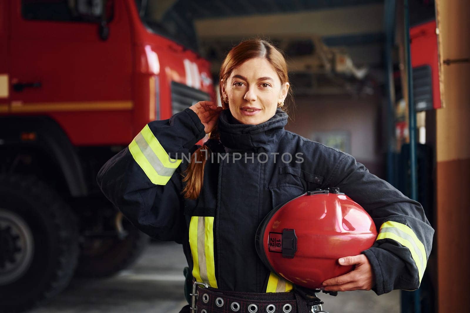 Holds red hat in hands. Female firefighter in protective uniform standing near truck.