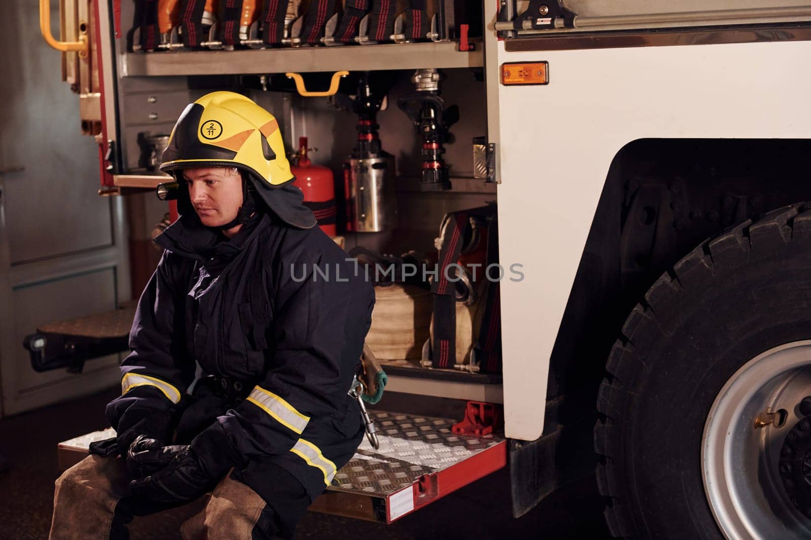 Sits on truck. Male firefighter in protective uniform. Feels tired.