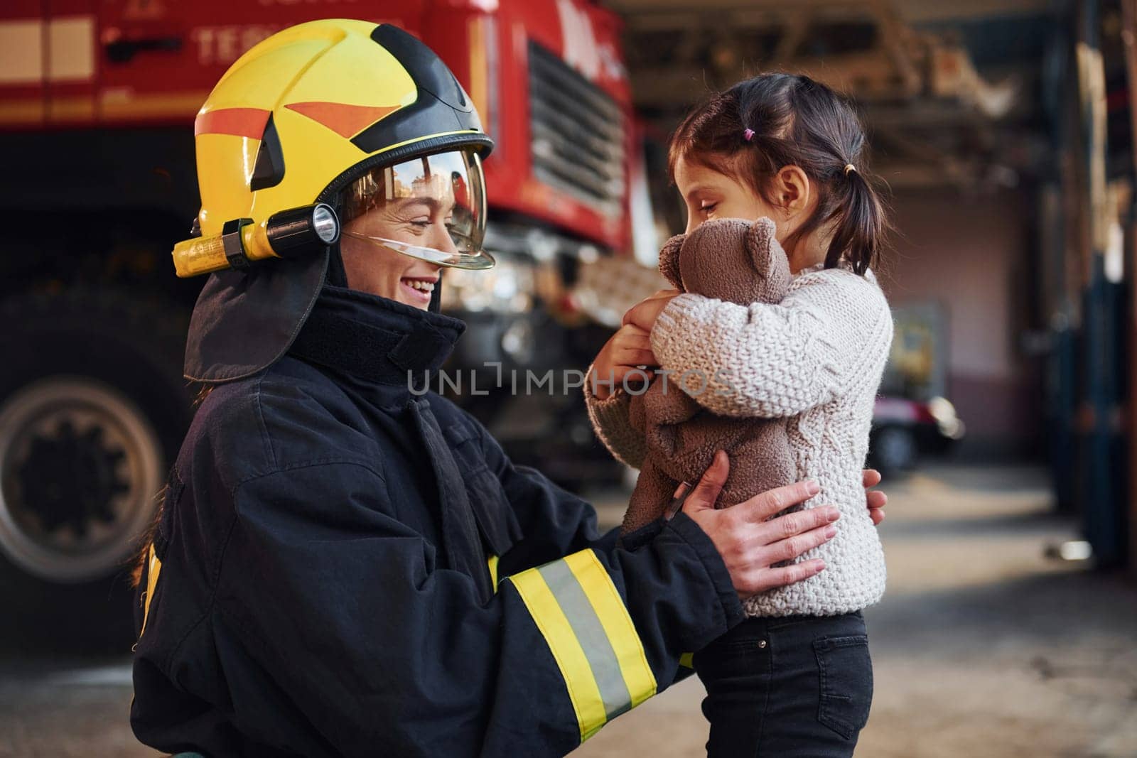 Happy little girl is with female firefighter in protective uniform.