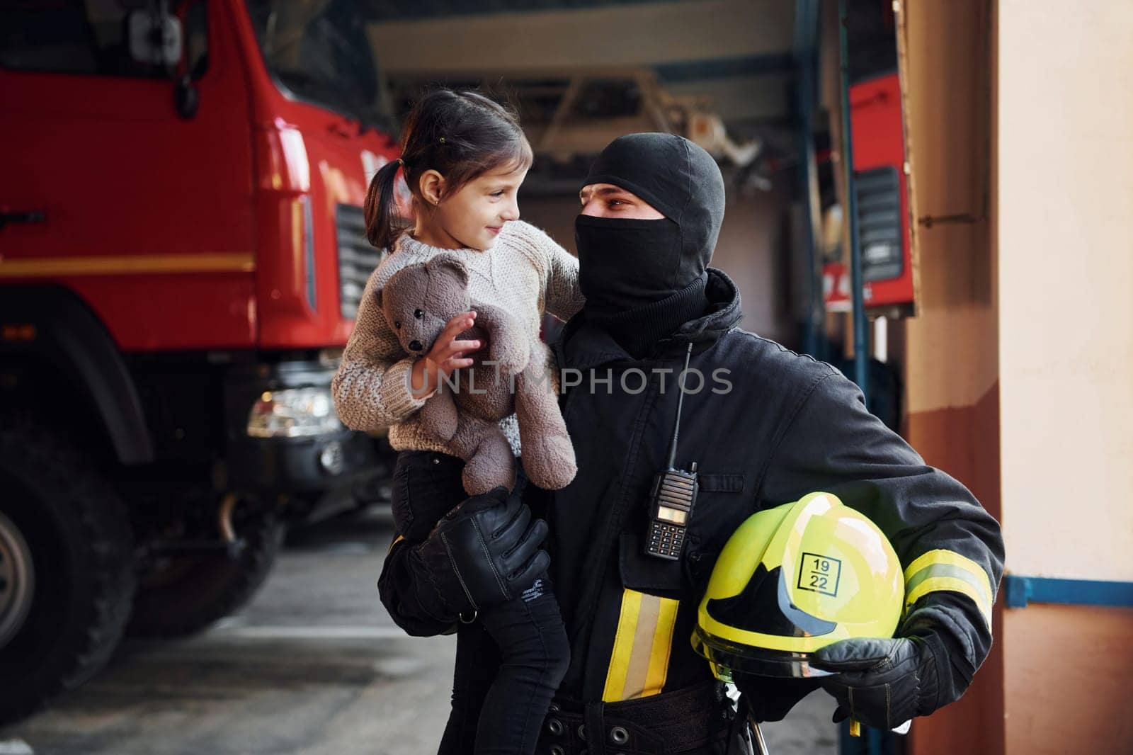 Holds teddy bear. Happy little girl is with male firefighter in protective uniform.