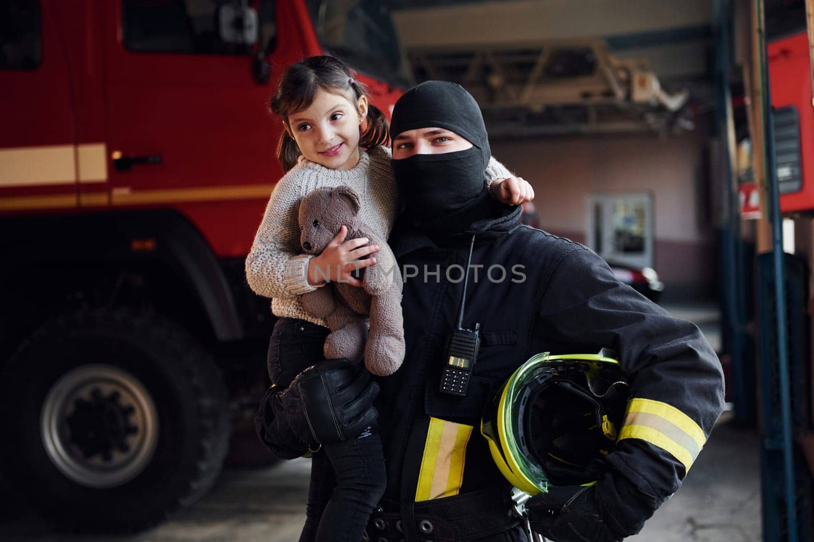 Holds teddy bear. Happy little girl is with male firefighter in protective uniform.