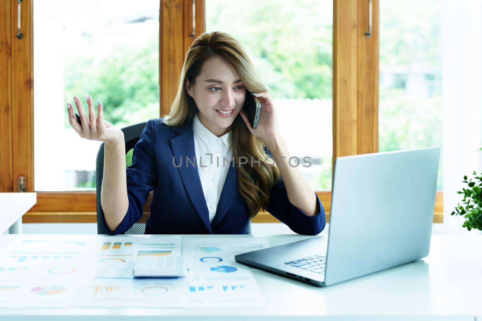Portrait of a young Asian woman showing a smiling face as she uses her phone, computer and financial documents on her desk in the early morning hours.