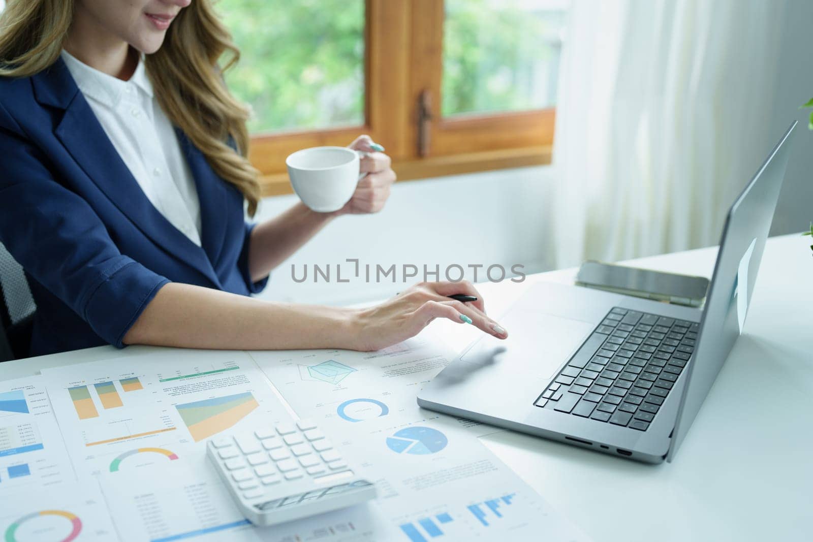 Portrait of an Asian business woman drinking coffee while working with a computer and financial statements documents on her desk by Manastrong