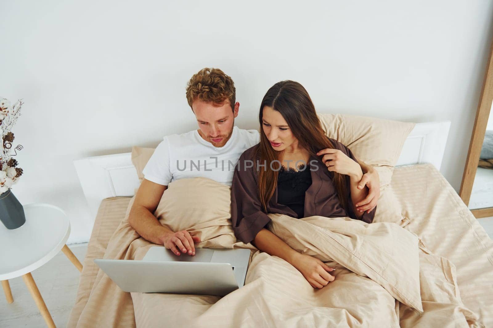 Couple lying down on bed with laptop. Interior and design of beautiful modern bedroom at daytime.
