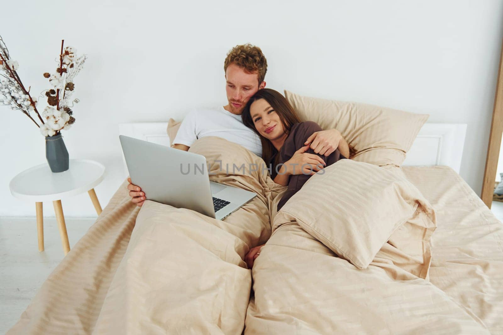 Couple lying down on bed with laptop. Interior and design of beautiful modern bedroom at daytime.