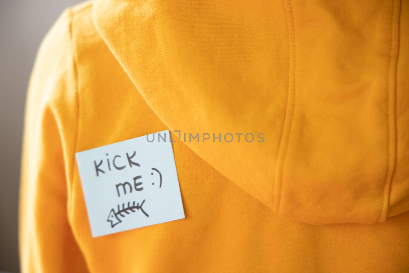 Boy in yellow hoodie with a sticky note on his back, saying kick me.