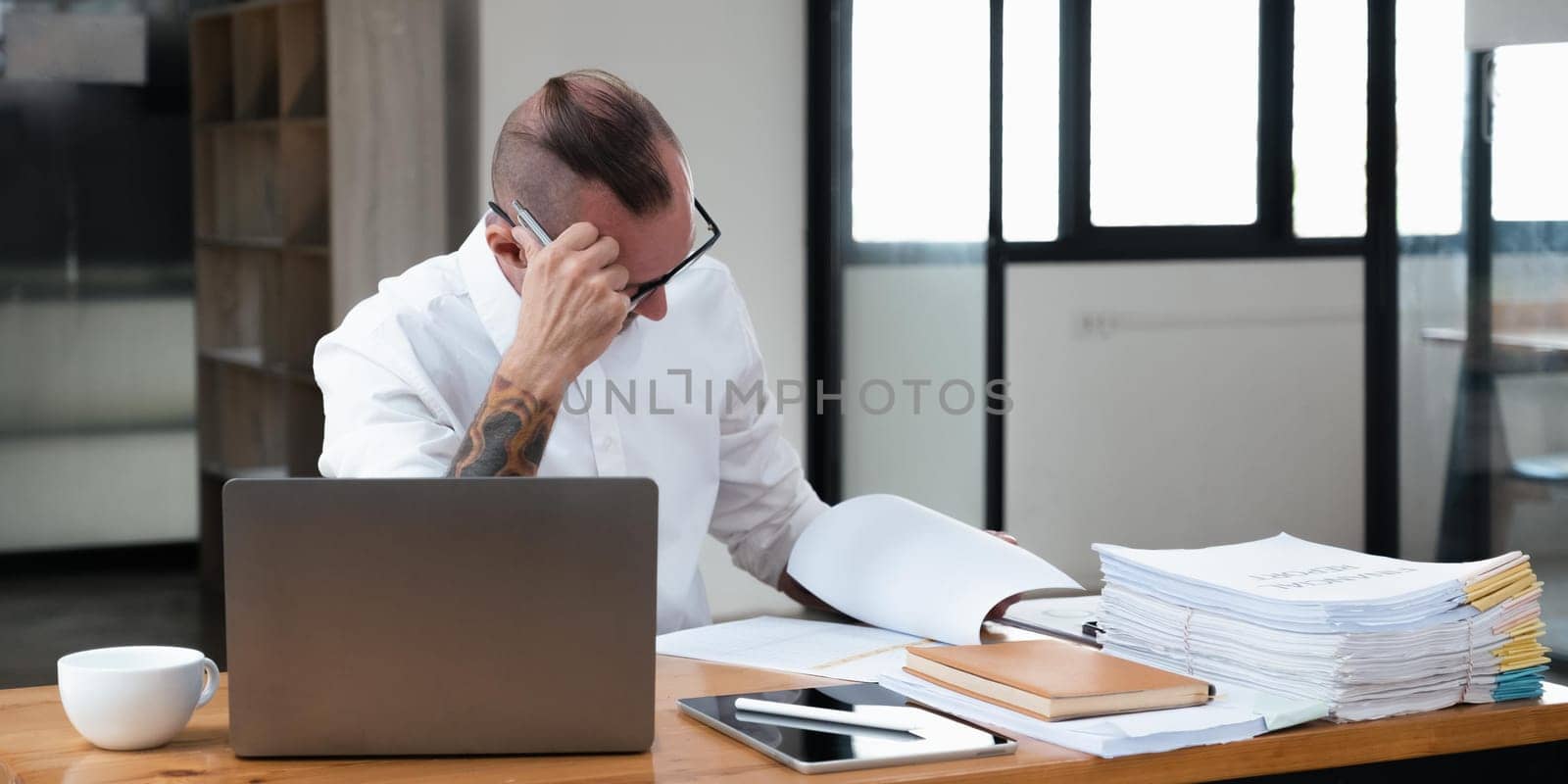 Serious business man using laptop sitting at the table in a home office, looking at the paper, communicating online, writing emails, distantly working on computer.