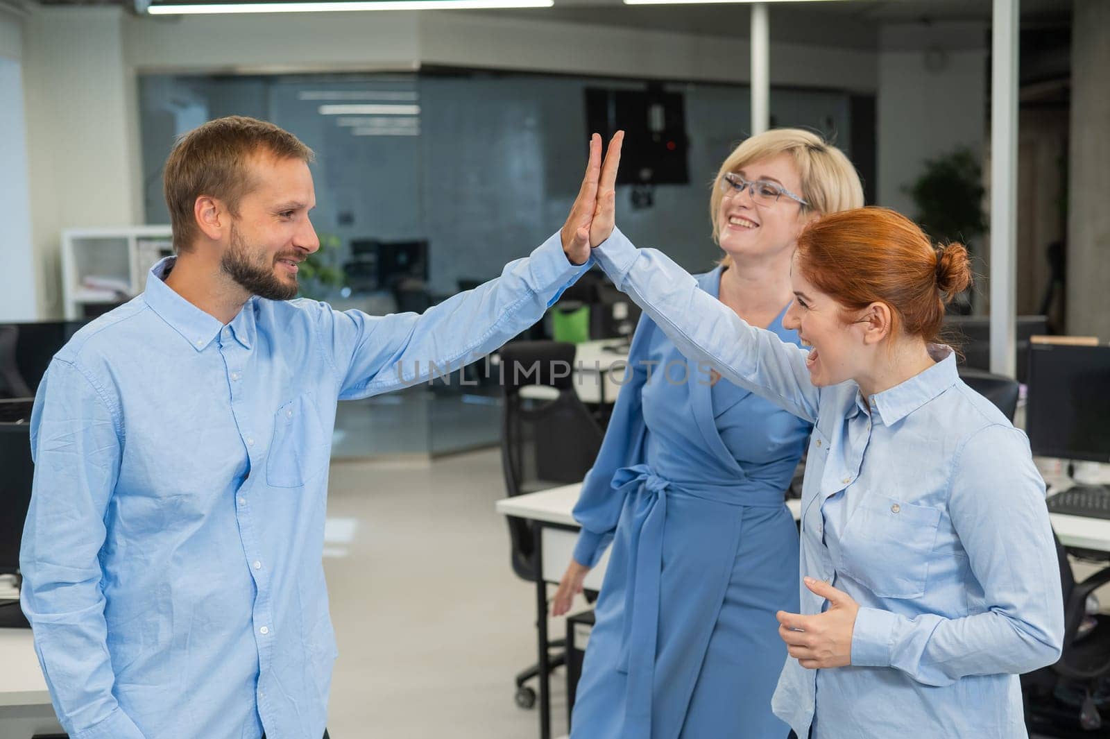 Colleagues give a high five in the office. Red-haired woman, blonde and bearded man in a denim shirt in the office