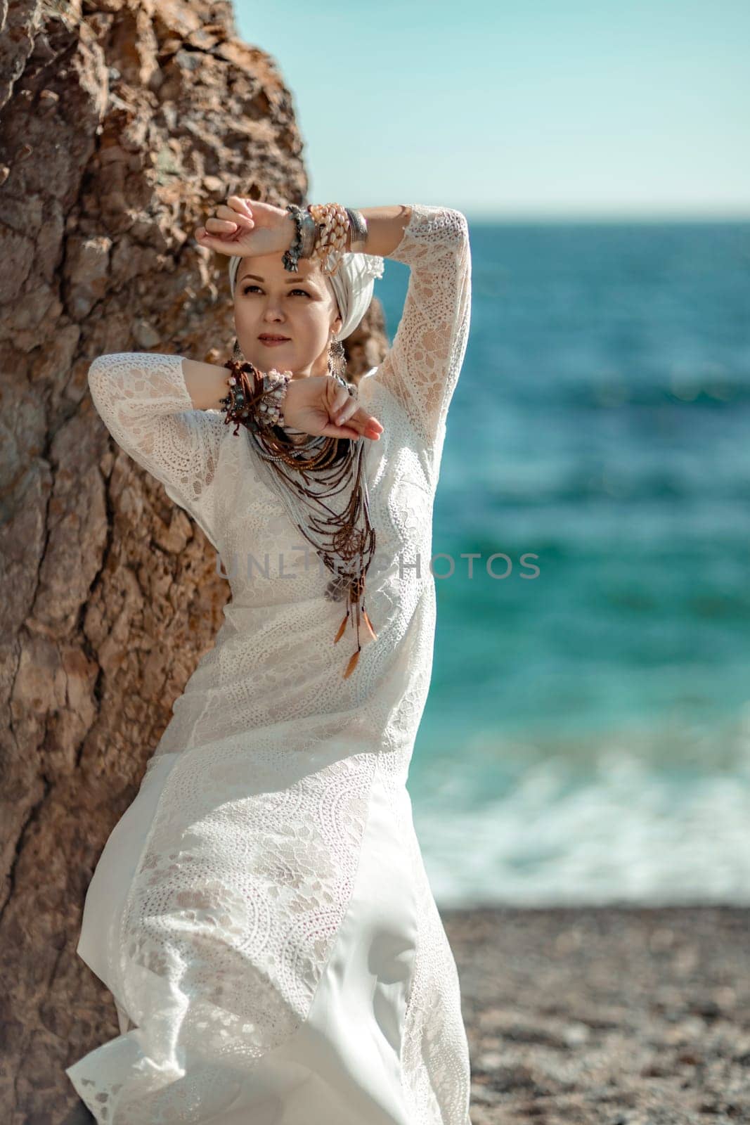 Woman white dress sea stones rocks.Middle-aged woman looks good with blond hair, boho style in a white long dress on beach jewelry around her neck and arms. by Matiunina