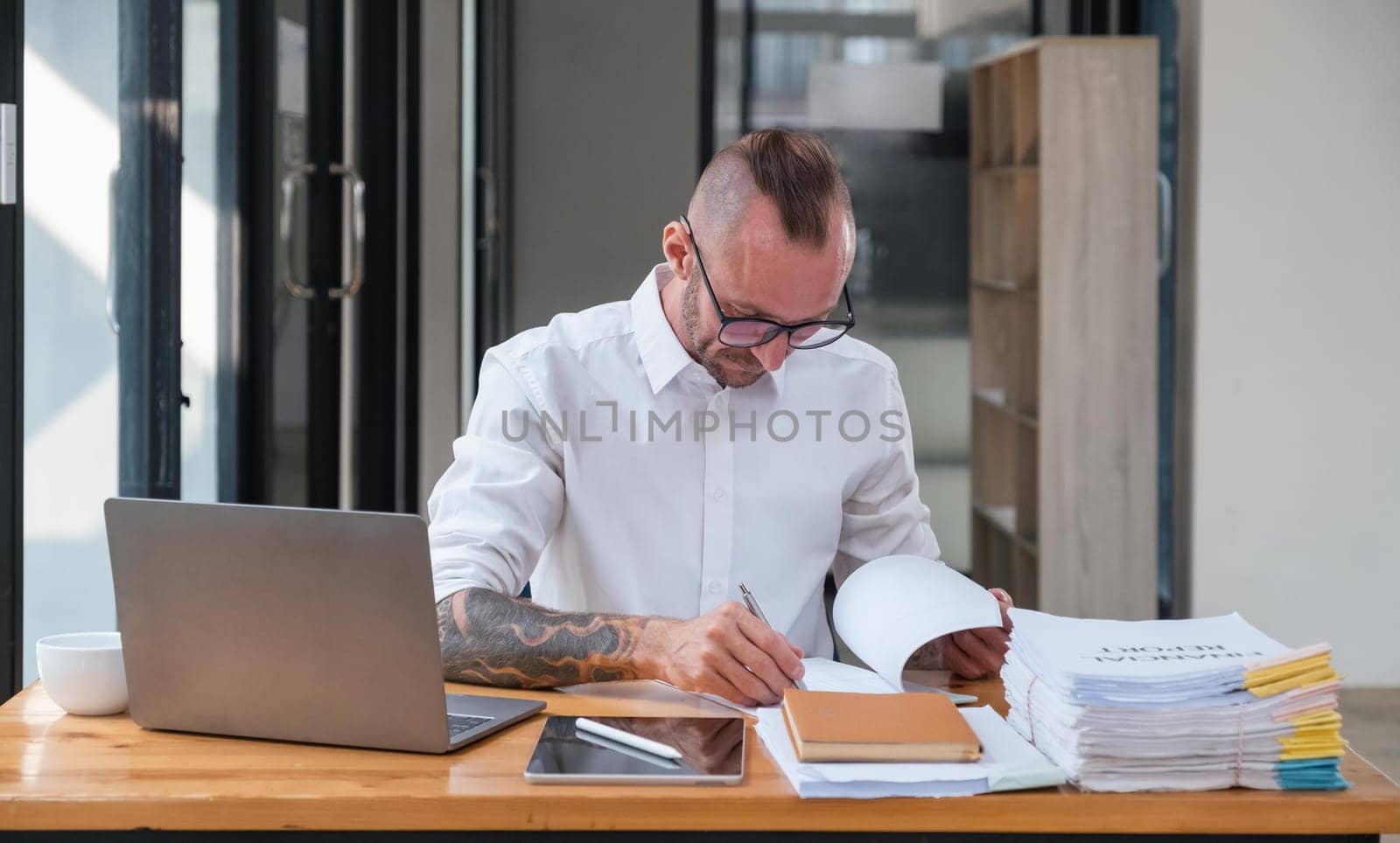 Serious business man using laptop sitting at the table in a home office, looking at the paper, communicating online, writing emails, distantly working on computer.