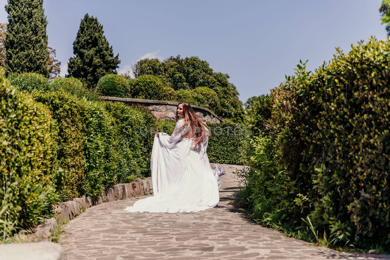 Brunette runs white dress park. A beautiful woman with long brown hair and a long white dress runs along the path along the beautiful bushes in the park, rear view.