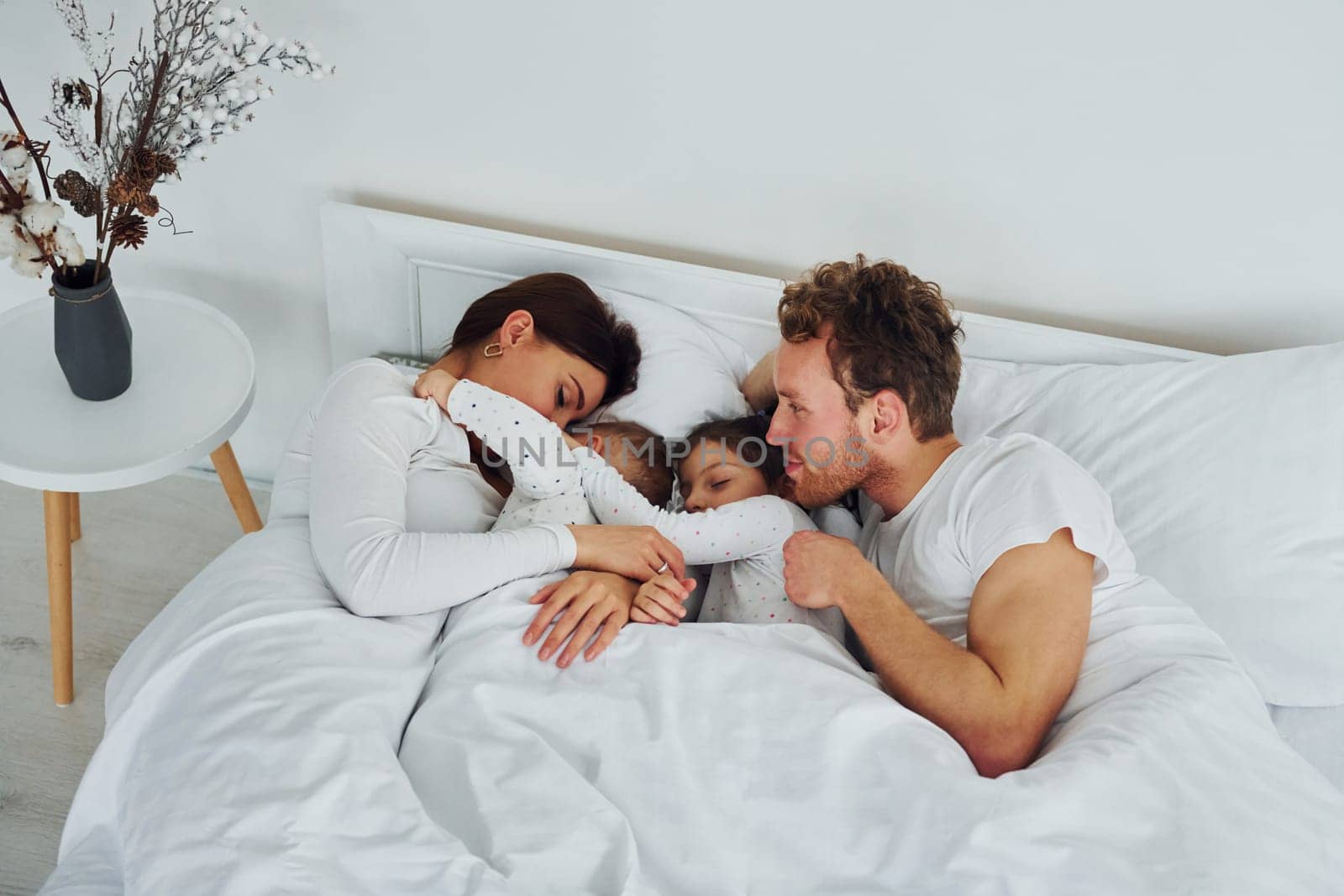 Woman and man lying down on bed with their children. Interior and design of beautiful modern bedroom at daytime.