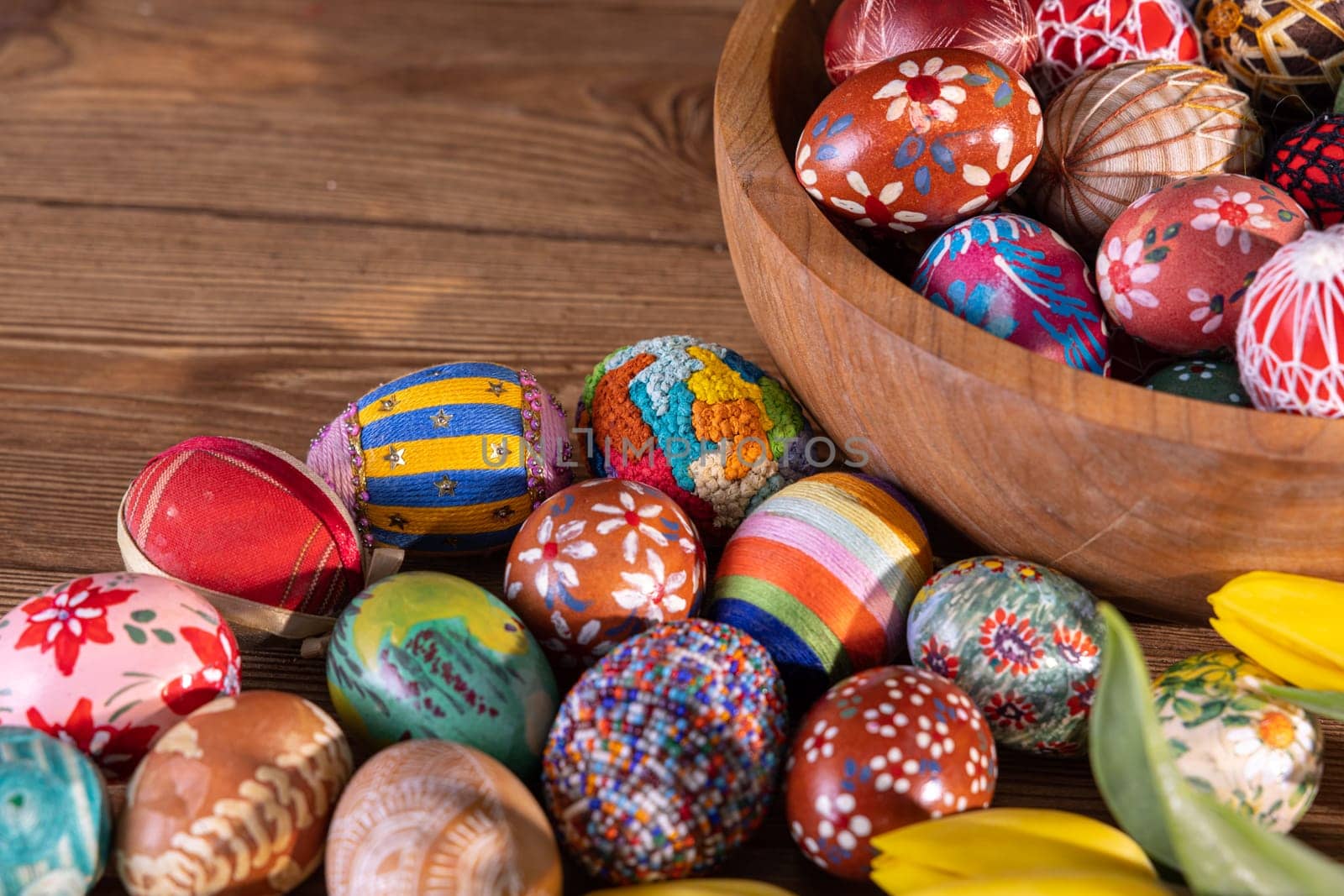 Intricately arranged Easter eggs on a wooden tabletop and in a wooden bowl. The Easter composition is completed with swishy yellow tulips.