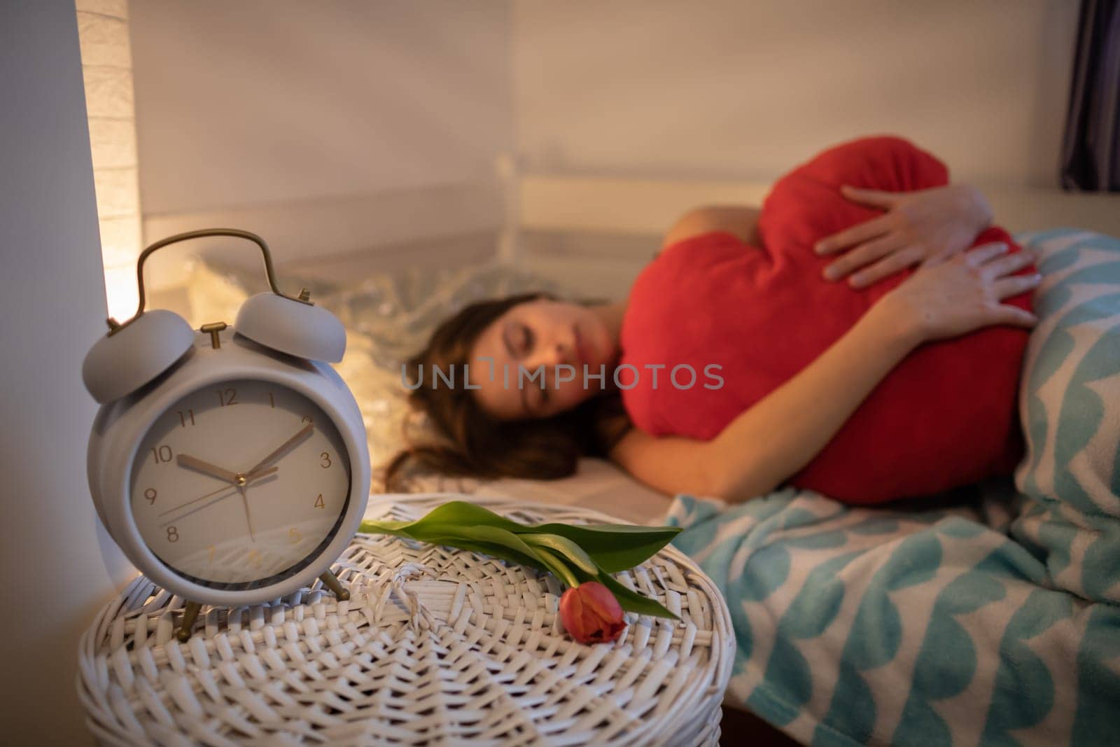 Girl in love hugs her heart-shaped pillow in red color. Bright bedroom with decorations. A large alarm clock stands on the bedside table.