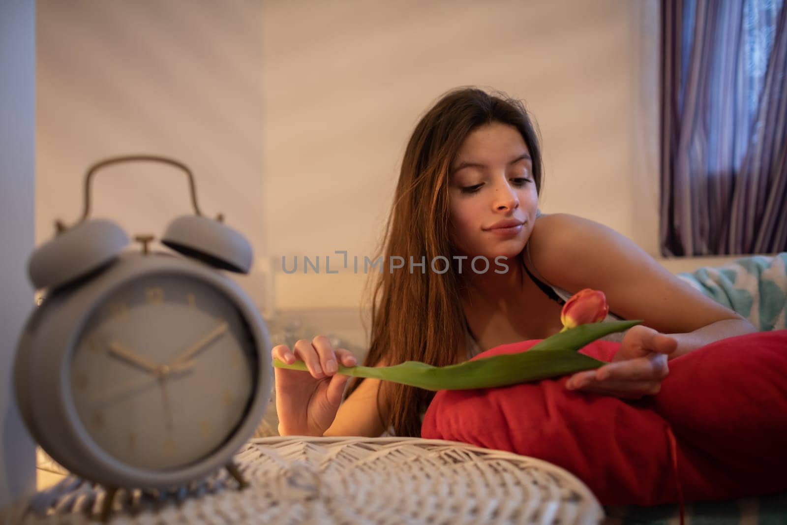 A woman lies in bed with a red pillow and holds a tulip. A large alarm clock stands on the bedside table.
