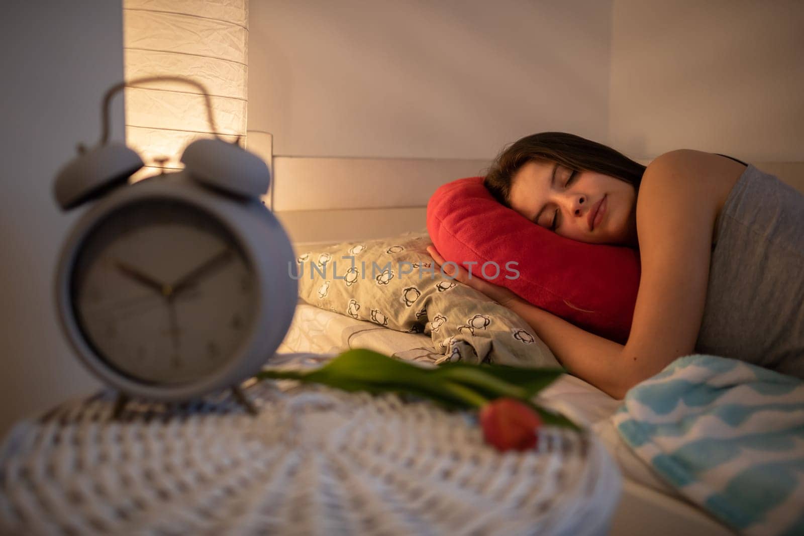 A young woman sleeps snuggled into her red pillow. The light of a lamp on the wall.