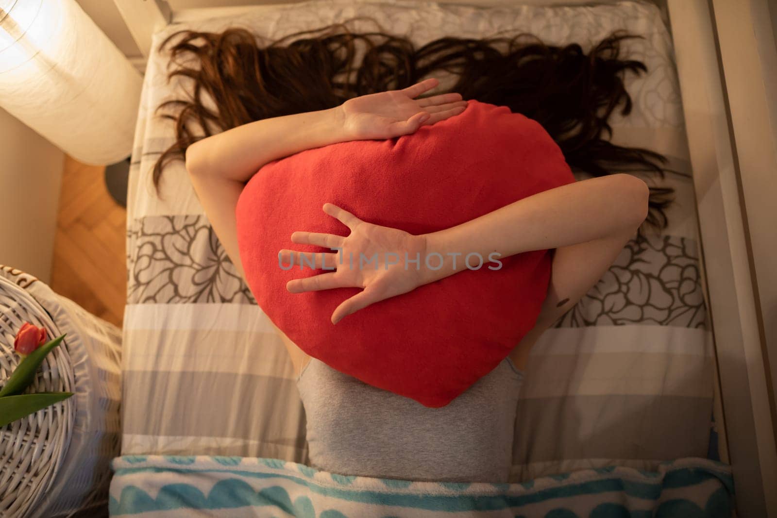 The red heart-shaped pillow is lying on the girls face. The light of a lamp on the wall.