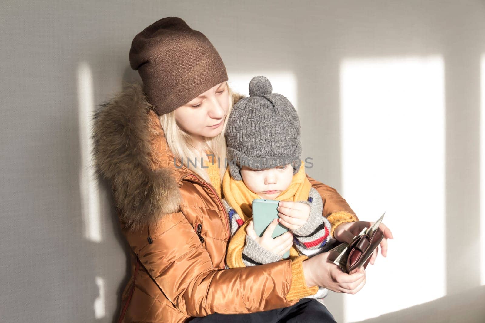 A mother with a small child in her arms is warmly dressed in outerwear, counting money in her purse. The concept of the economic crisis and large utility bills in homes. by Alla_Yurtayeva