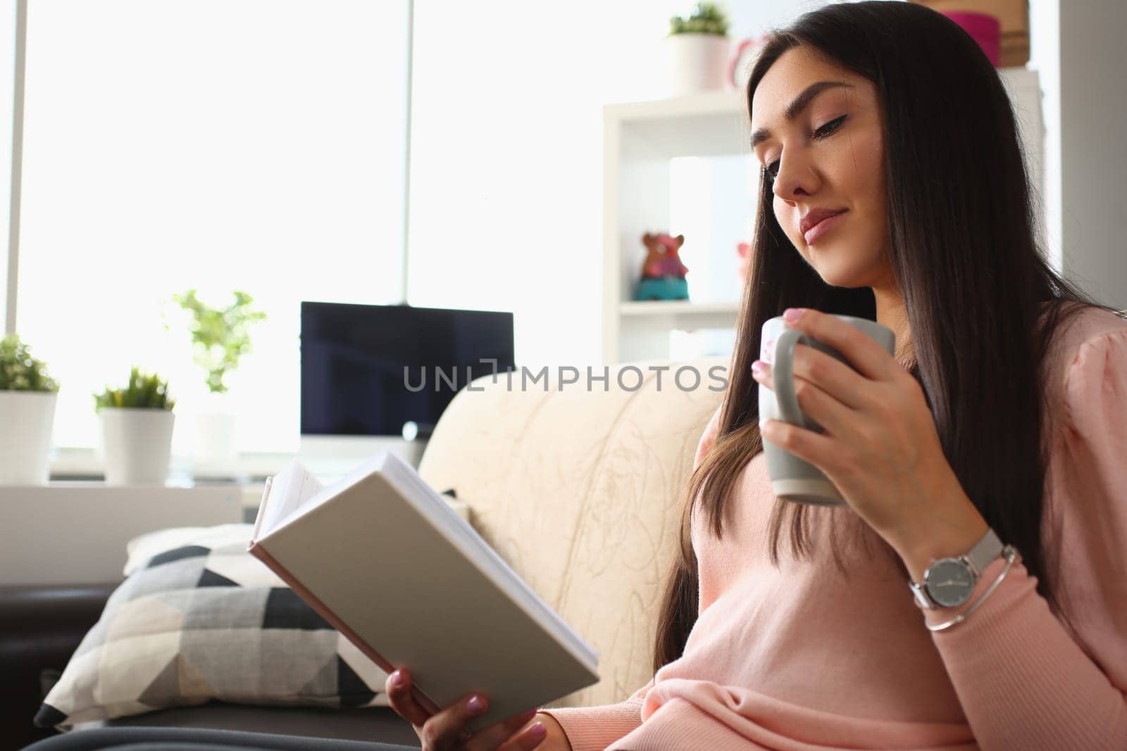 Young woman is reading book and holding cup in living room on sofa. Leisure and reading books concept